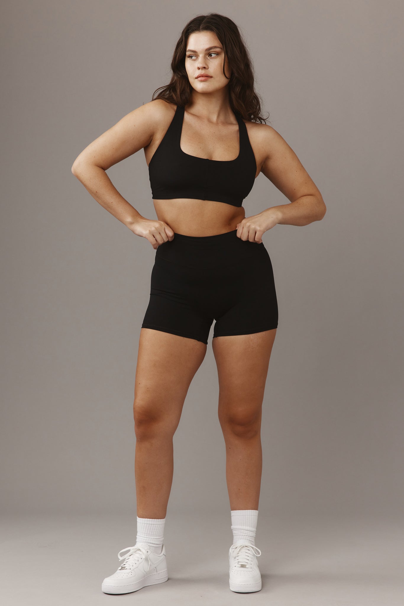 Woman modeling black sports bra and high-waist bike shorts, hands on waistband, white sneakers, neutral gray backdrop