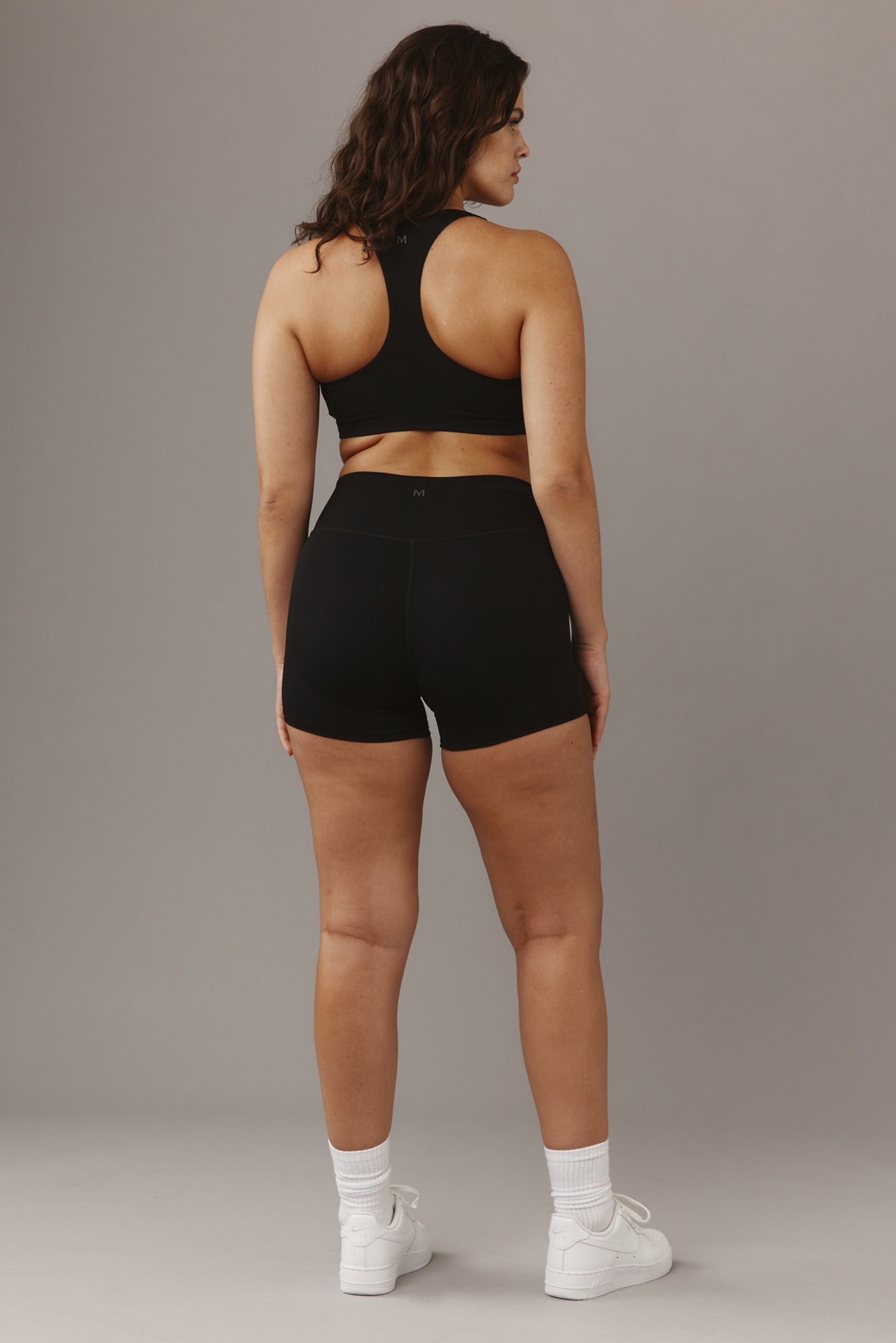Rear view of woman wearing black racerback sports bra and black high-waist shorts with M logo, white sneakers, gray studio background.