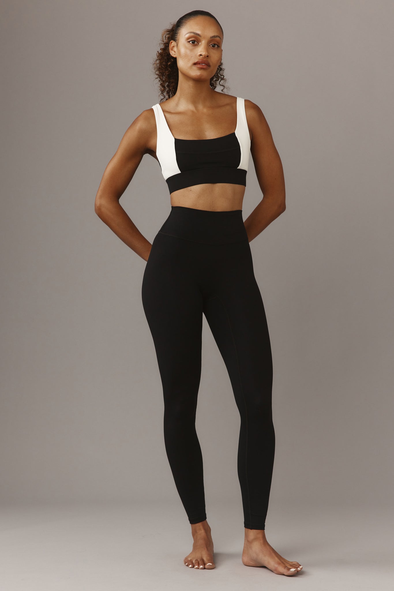Model standing in high-waist black leggings and black-and-white sports bra on neutral background, hands behind back.