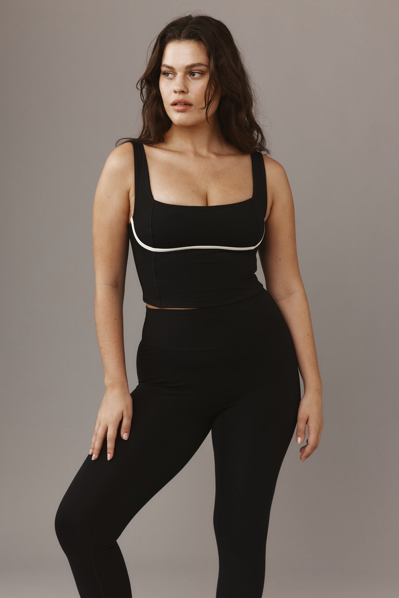 Woman in black cropped tank with white piping and high-waist black leggings, standing against a gray background, looking left.