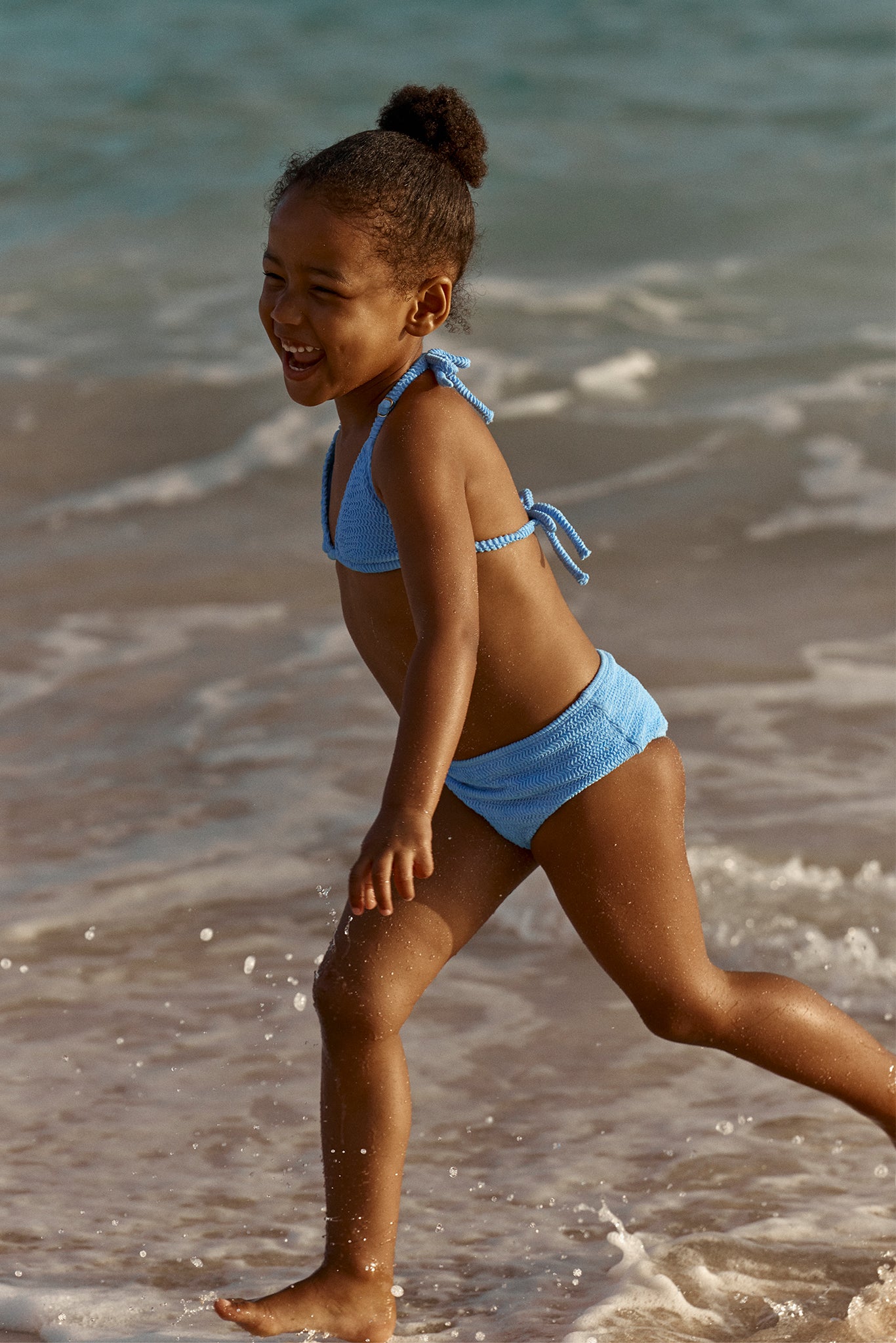 Young girl laughing as she runs along the shoreline in a blue bikini, water splashing around her feet