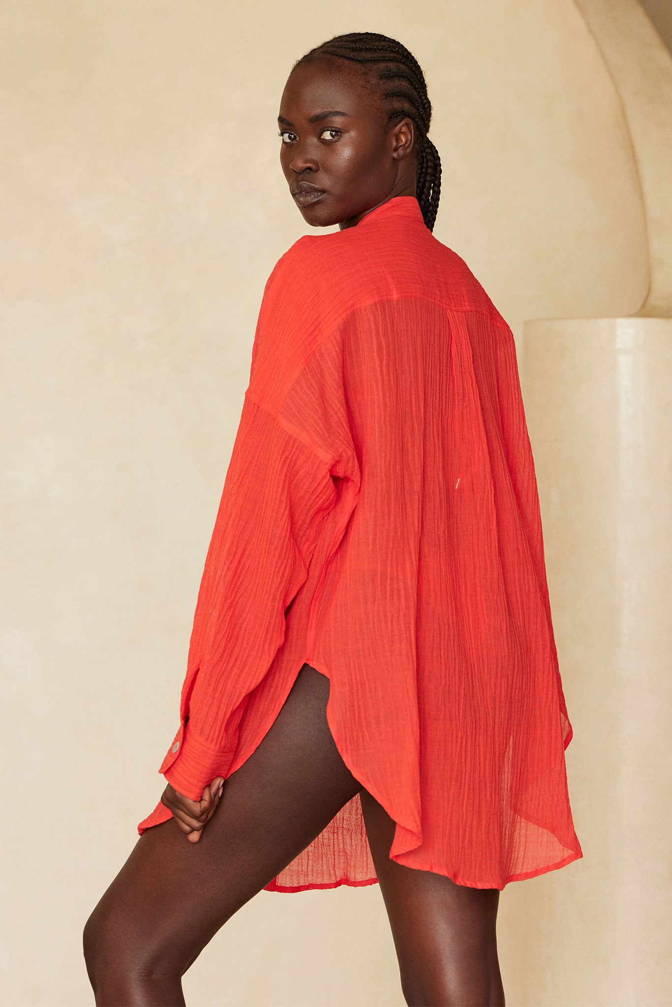 Model with braided hair looking over shoulder, wearing a sheer red oversized shirt against a beige backdrop.