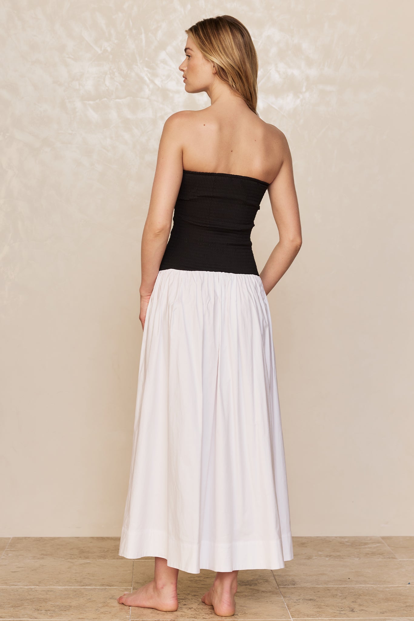 Back view of a woman wearing a black strapless top with a white ankle-length skirt standing barefoot on a tiled floor.