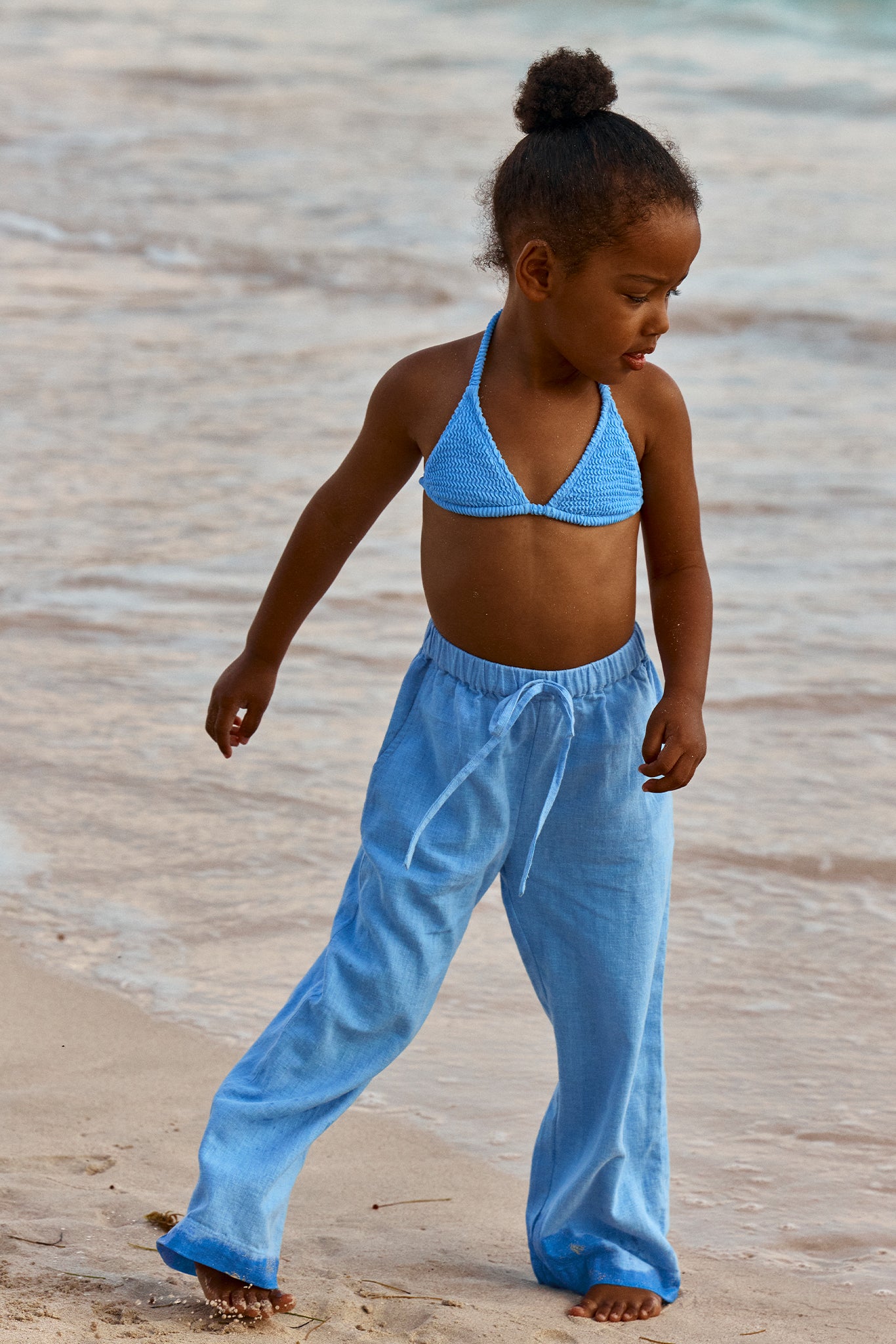 Young girl in blue bikini top and loose light-blue beach pants walking barefoot on wet sand