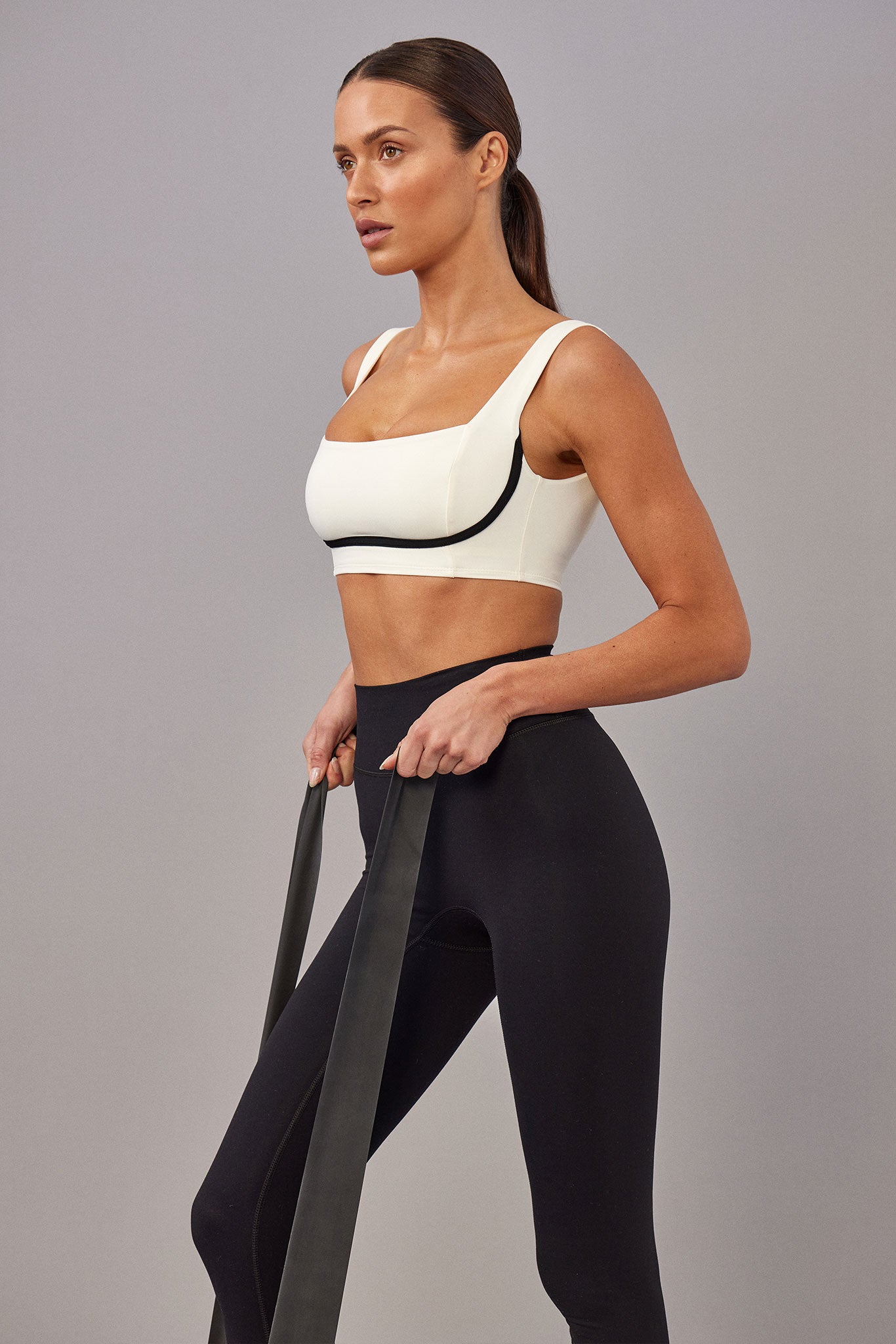 Model wearing cream sports bra and black leggings, standing while holding a long black resistance band.