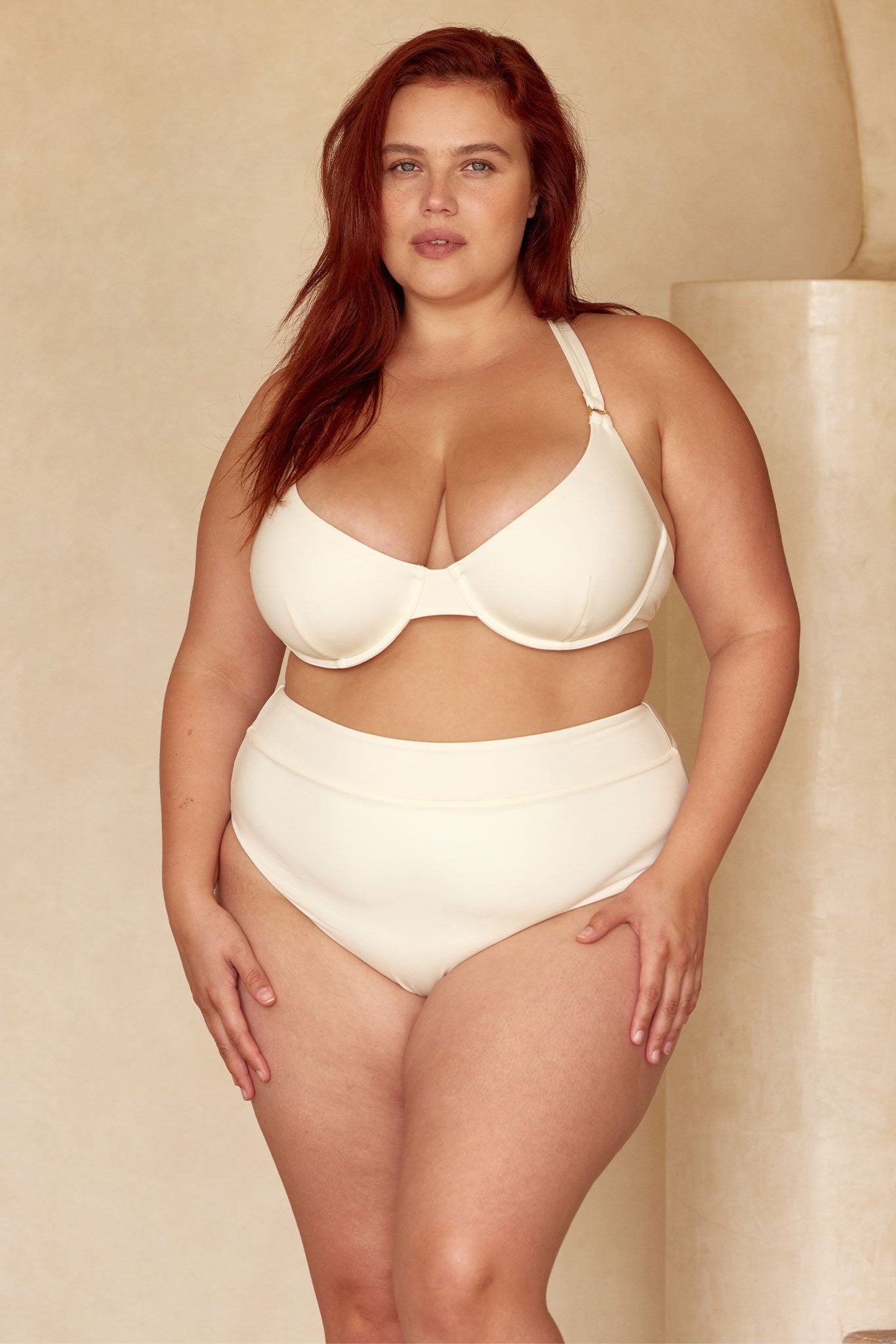 Plus-size woman standing in cream underwire bra and high-waist briefs against a neutral backdrop, hands resting at hips.
