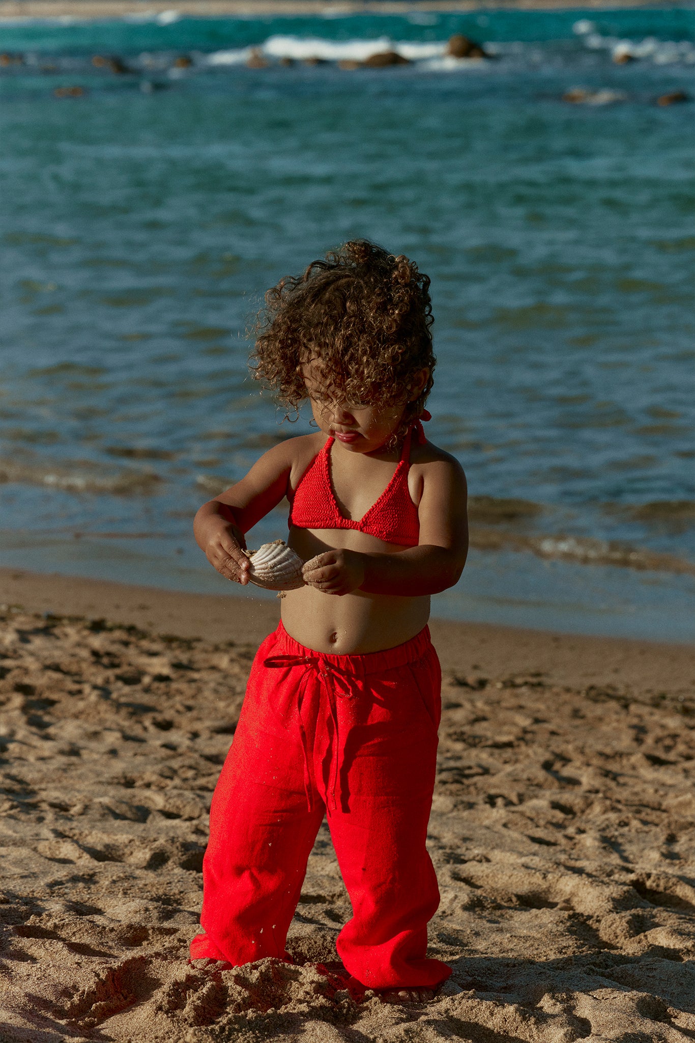 Toddler with curly hair in red bikini top and red pants on a sandy beach holding a seashell, ocean in background.