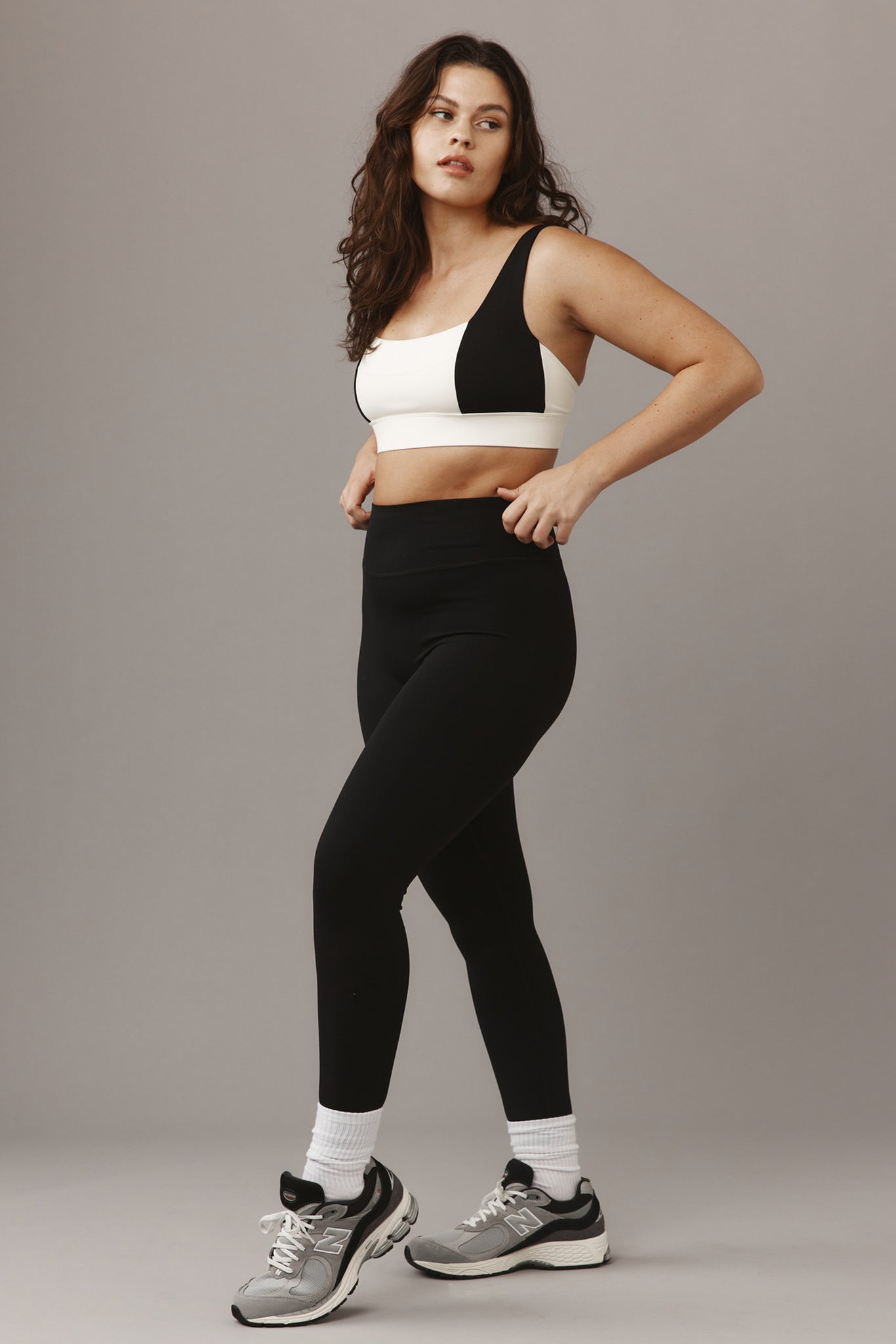 Woman adjusting waistband wearing white-and-black sports bra, high-waist black leggings and grey sneakers on neutral background