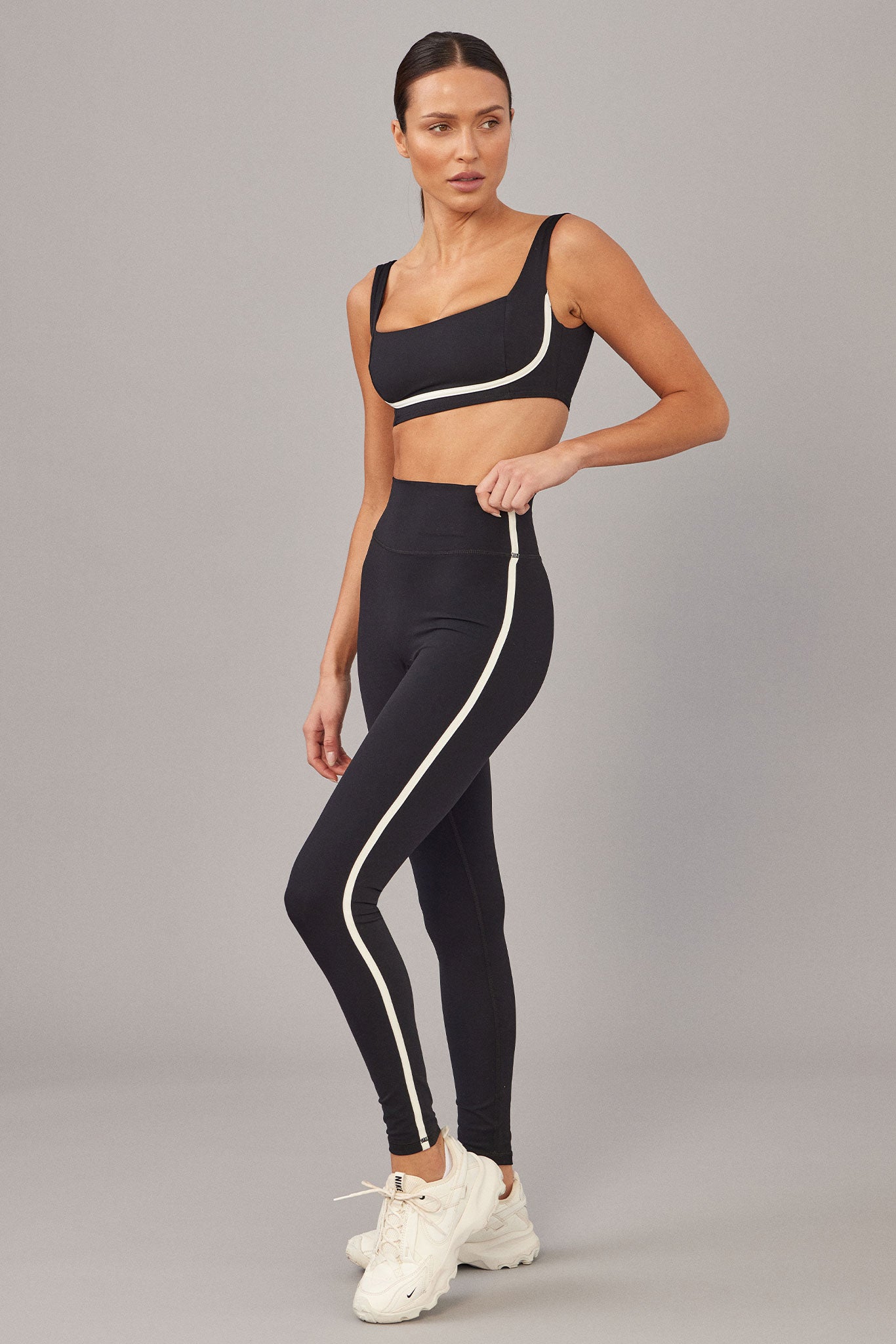 Model wearing black sports bra and high-waist leggings with white trim and white athletic sneakers.