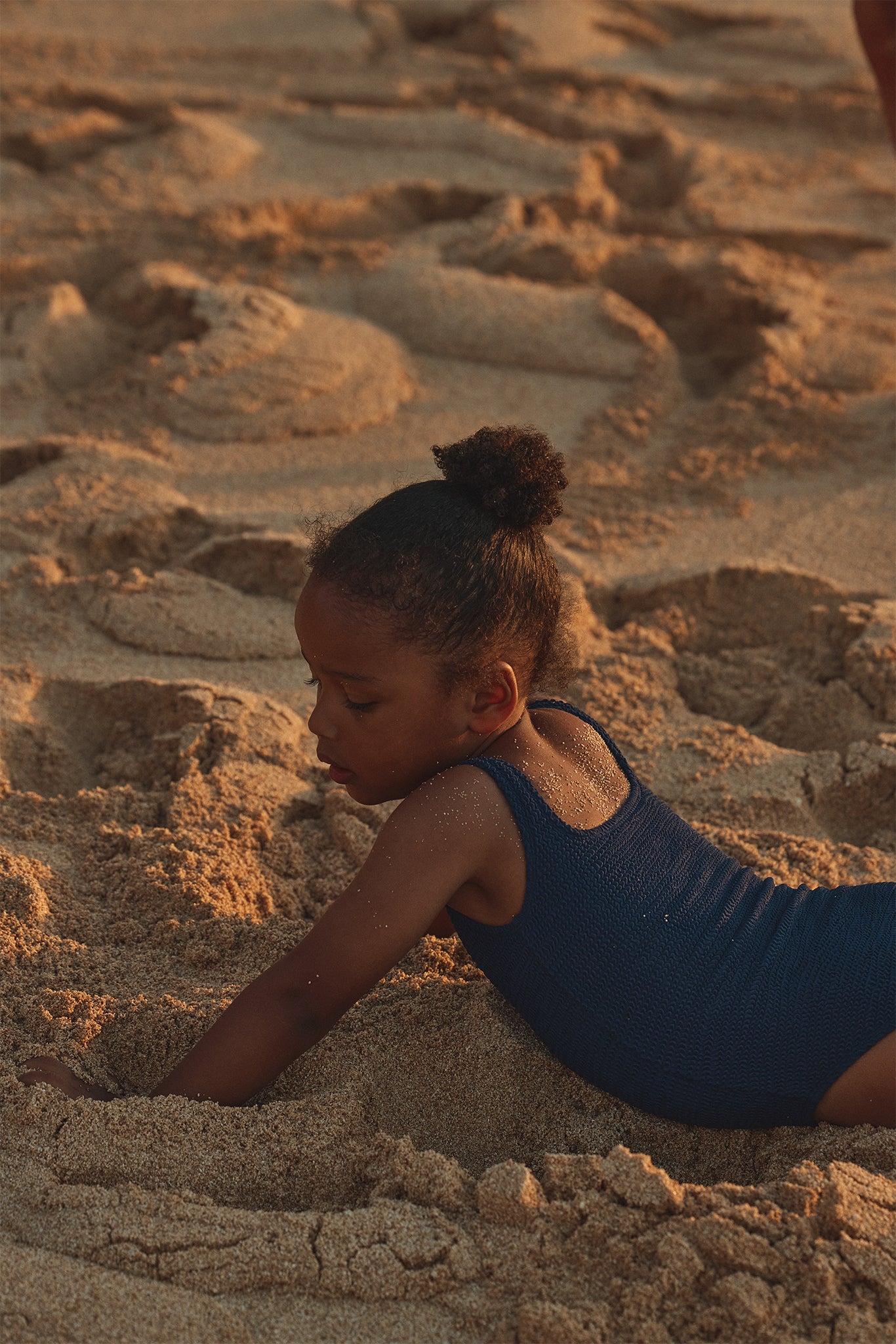 Child in a navy one-piece swimsuit lying on a sandy beach, reaching into the sand in warm sunset light.