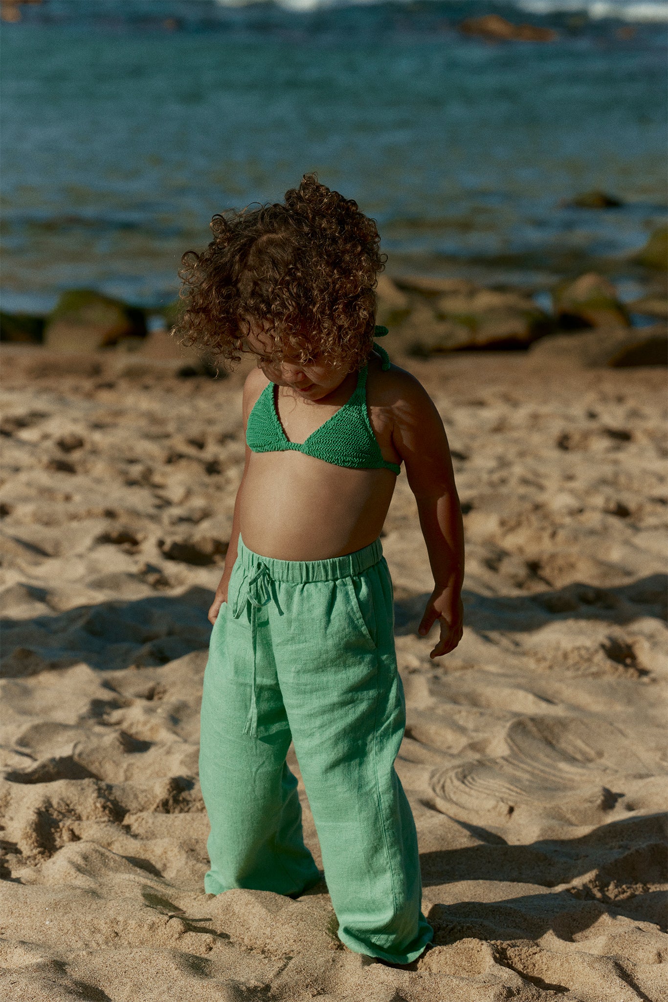 Young child with curly hair wearing a green knit bikini top and loose mint pants standing on sunlit sandy beach, looking down
