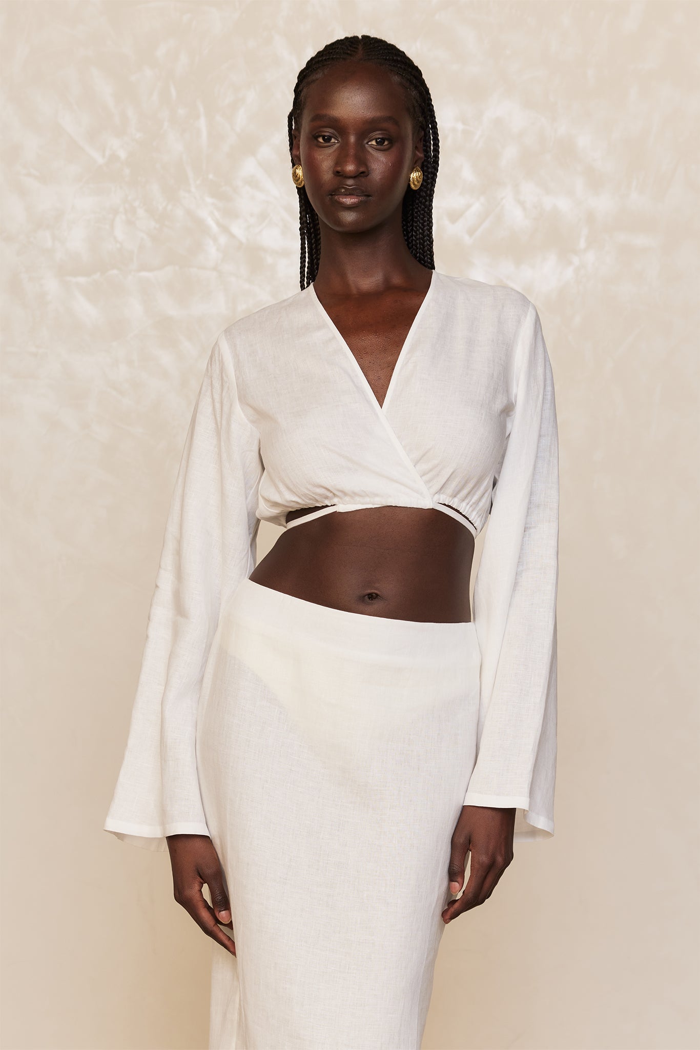 Dark-skinned model wearing white wrap crop top and high-waist linen skirt, braided hair and gold earrings, neutral backdrop.