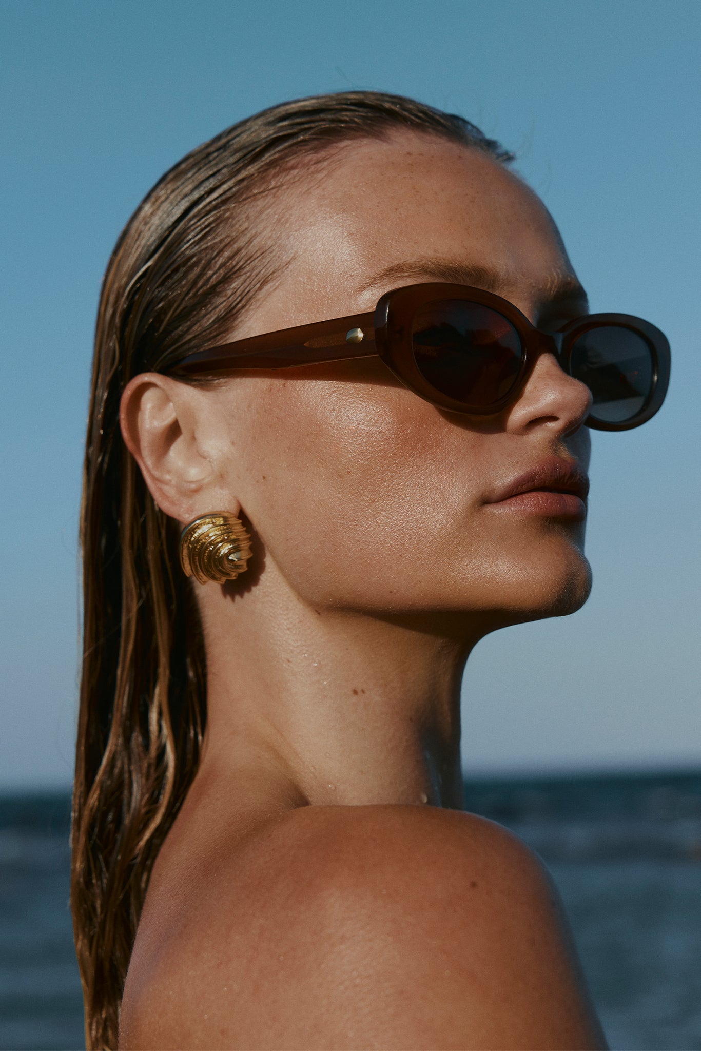 Close-up profile of a woman with wet hair wearing brown oval sunglasses and gold shell earrings, sea in background.
