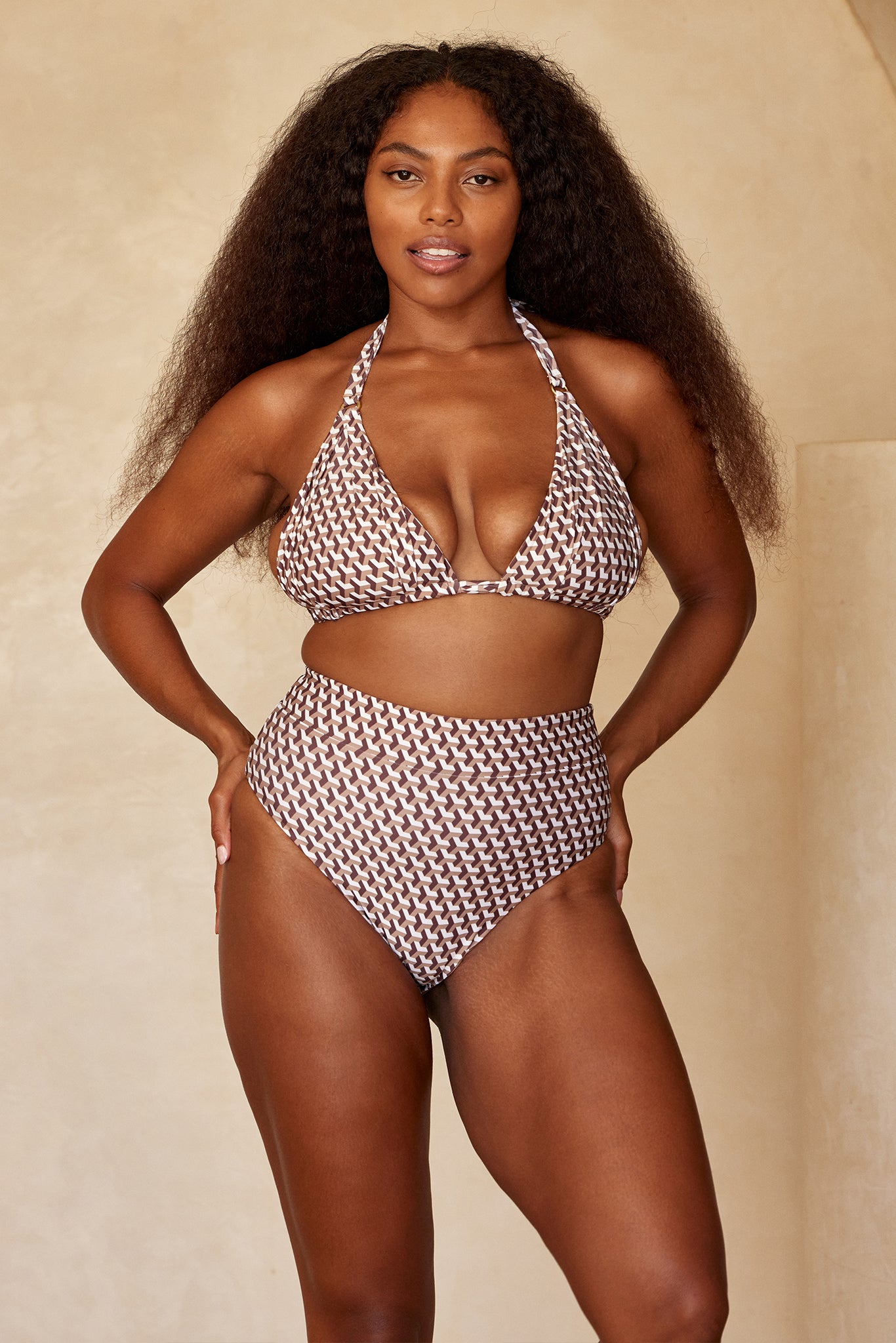 Model wearing brown-and-white geometric halter bikini top and high-waist bottoms, hands on hips, beige studio background.