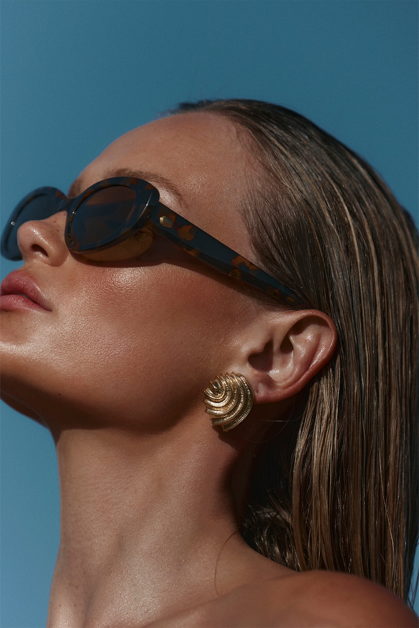 Close-up profile of a woman wearing tortoiseshell sunglasses and a large gold spiral earring against blue sky.