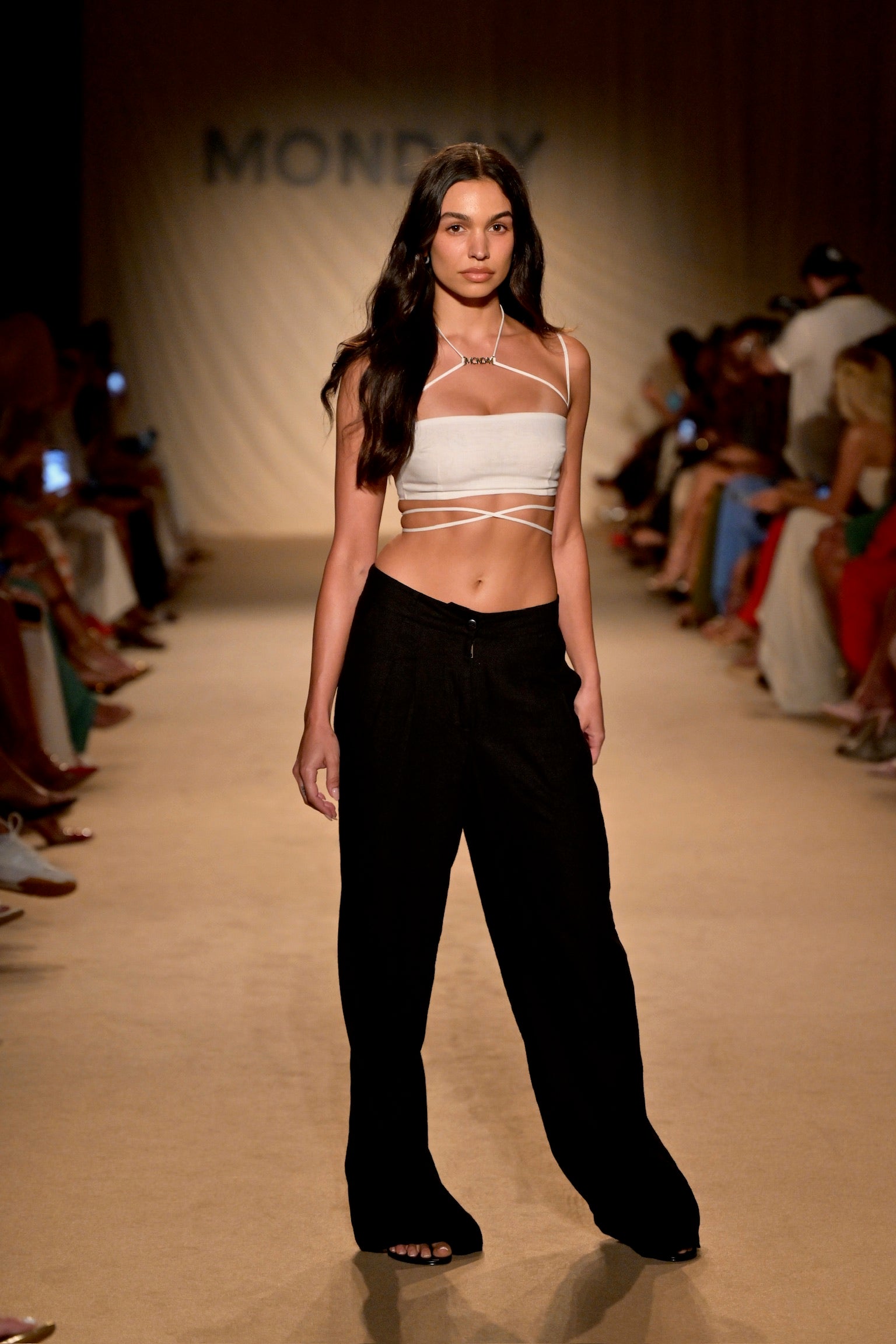 Model in white wrap crop top and black wide-leg pants walking the runway with backdrop reading MONDAY.