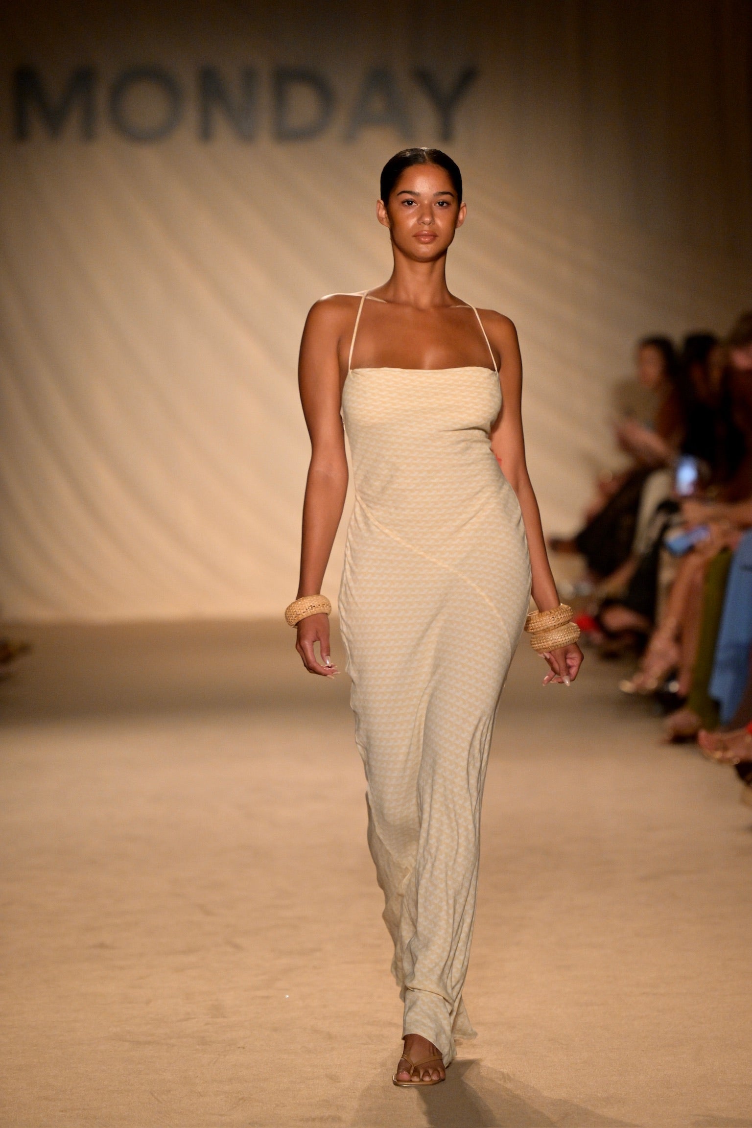 Model walking down runway wearing a beige strappy slip dress and woven bracelets, runway backdrop reads MONDAY.