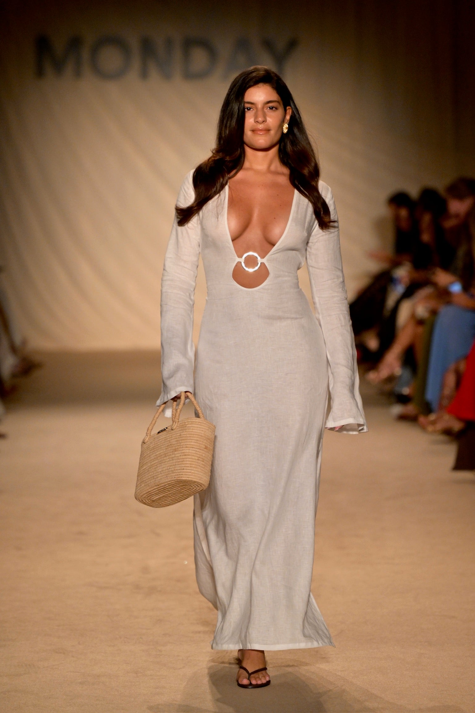 Runway model in long white dress with deep V and circular cutout, carrying woven straw bag; backdrop reads MONDAY