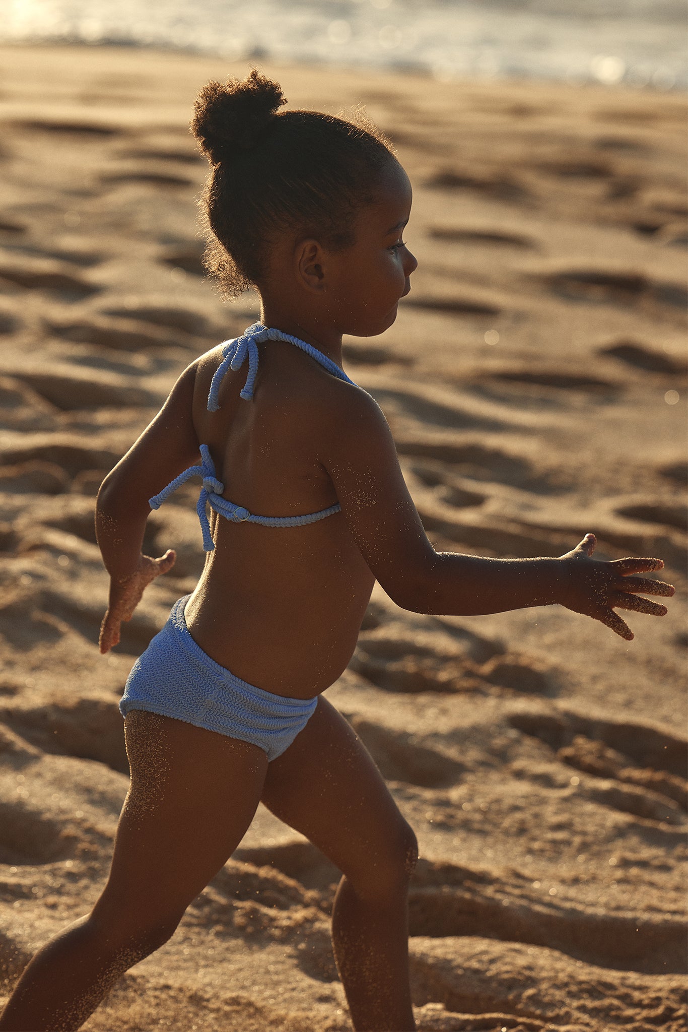 Small child in a blue knit bikini running along a sandy beach at sunset, side profile with sand on skin