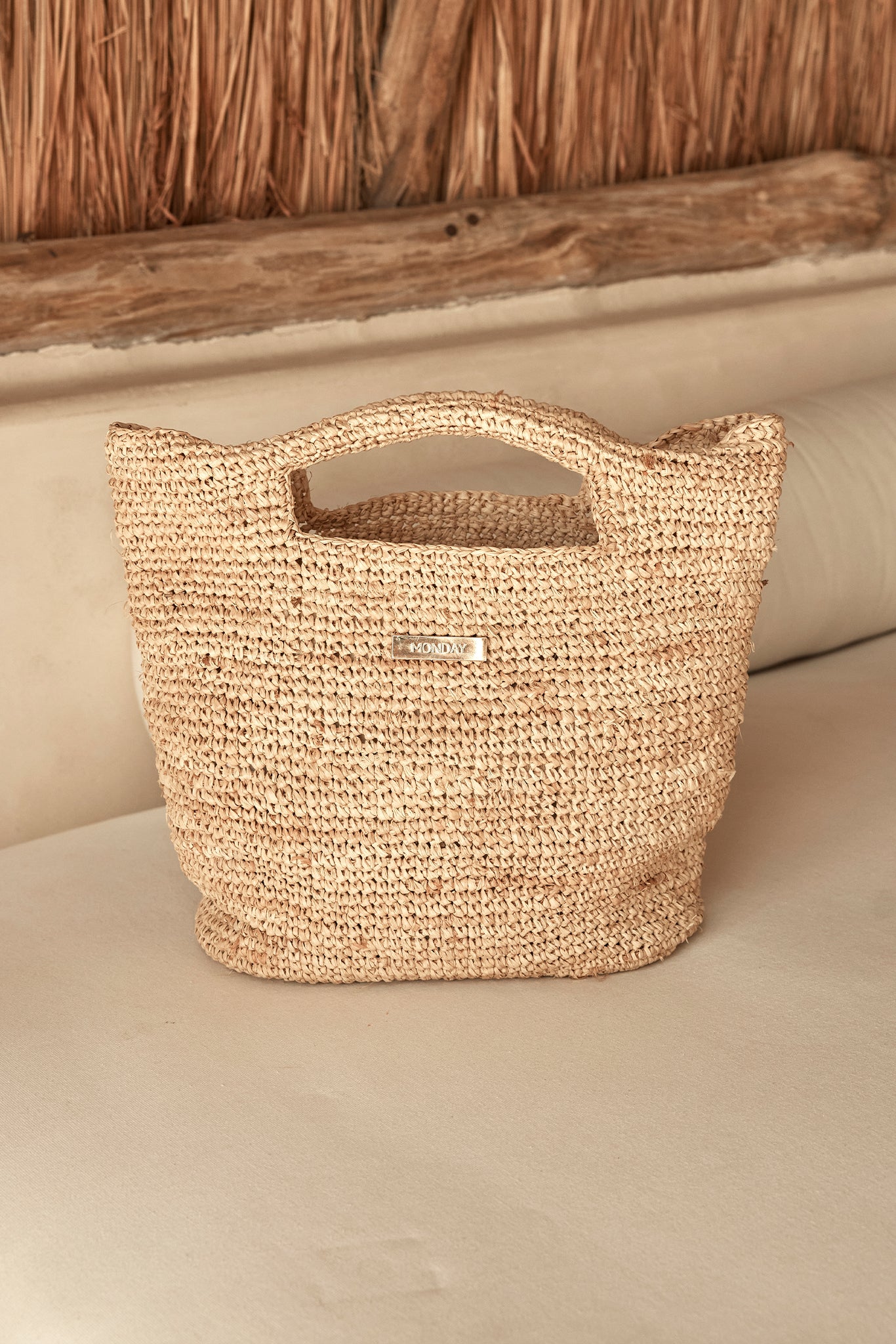 Handwoven straw tote with cutout handles and a metal label reading MONDAY, resting on a cream sofa.