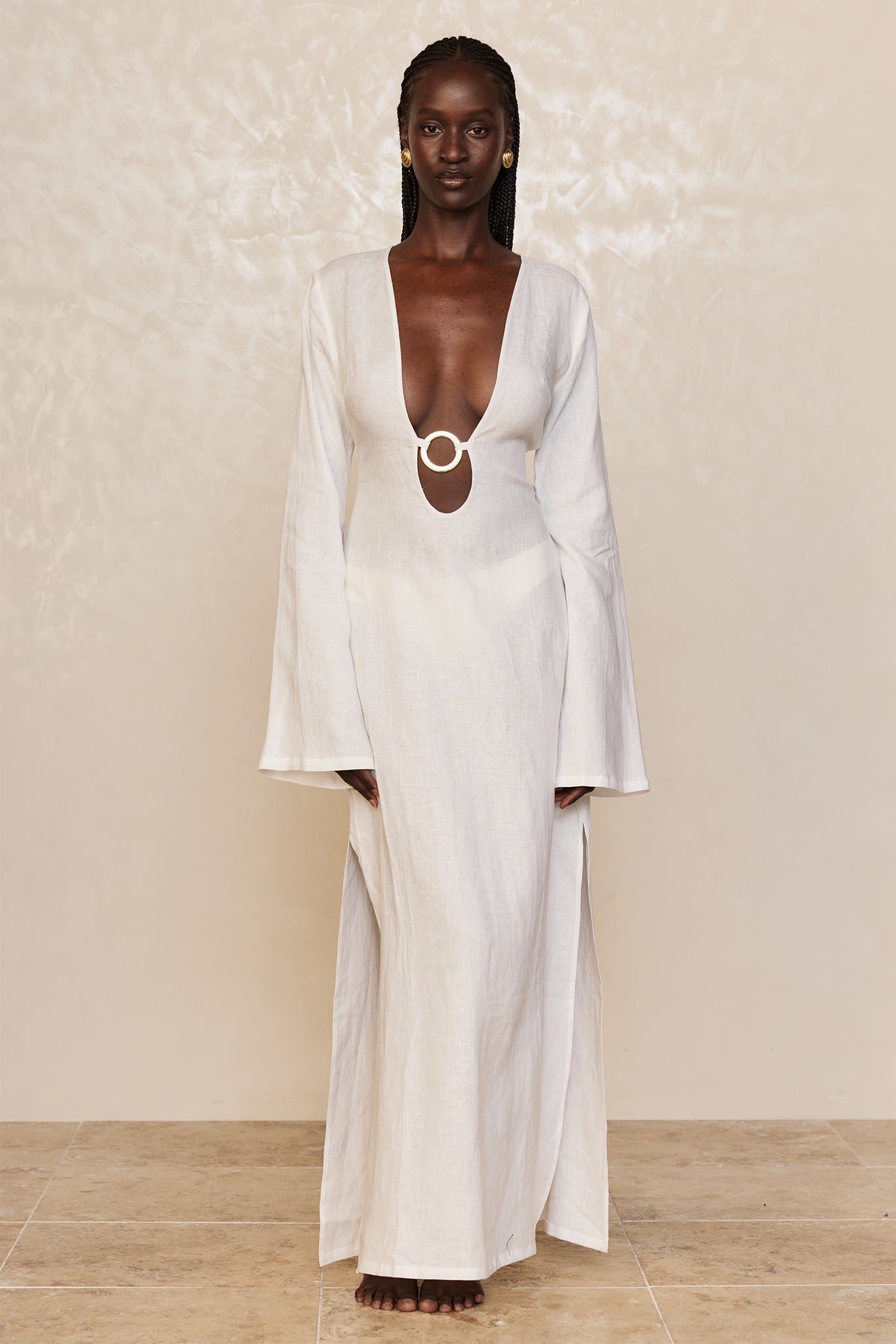Woman standing barefoot, facing camera, wearing long white linen dress with deep V neckline and circular ring detail at front.