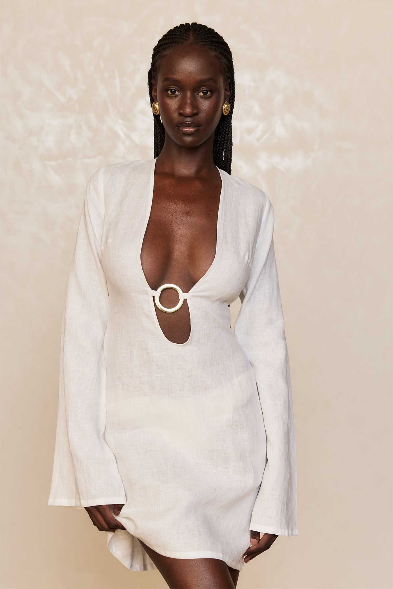 Dark-skinned model with braided hair wearing a white linen mini dress with deep V and gold ring detail at the waist, wearing gold disc earrings.