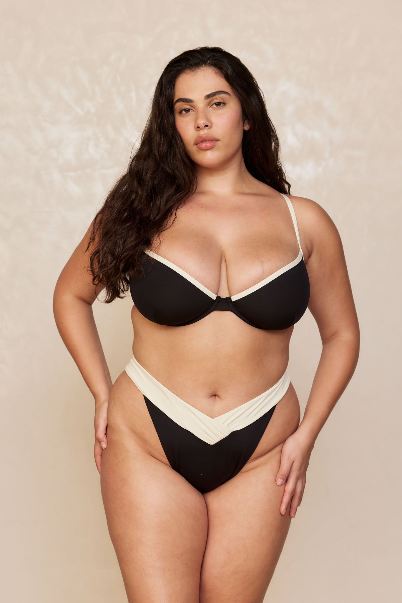 Plus-size model wearing black bikini with cream trim and V-front high-cut bottoms, standing against neutral backdrop.