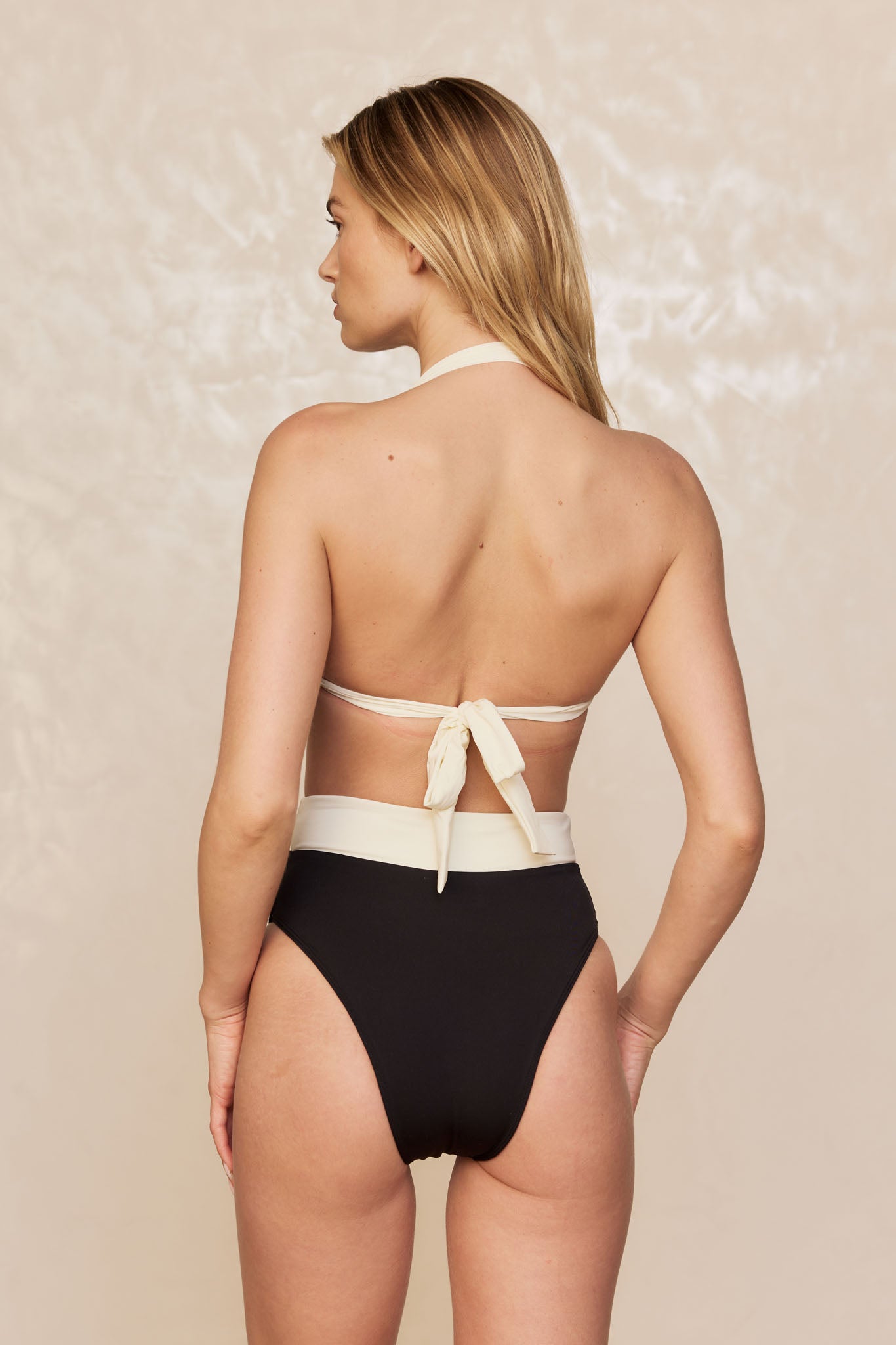 Back view of woman in cream halter-top and black high-waist bikini bottoms with bow tied at mid-back.