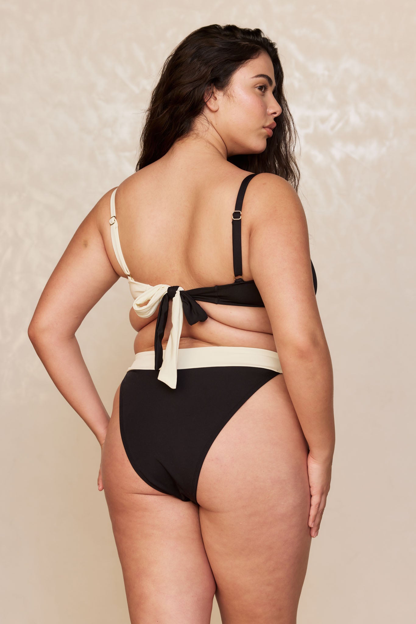 Plus-size woman seen from back wearing a black-and-cream two-piece bikini with tied bow at back, neutral studio background.