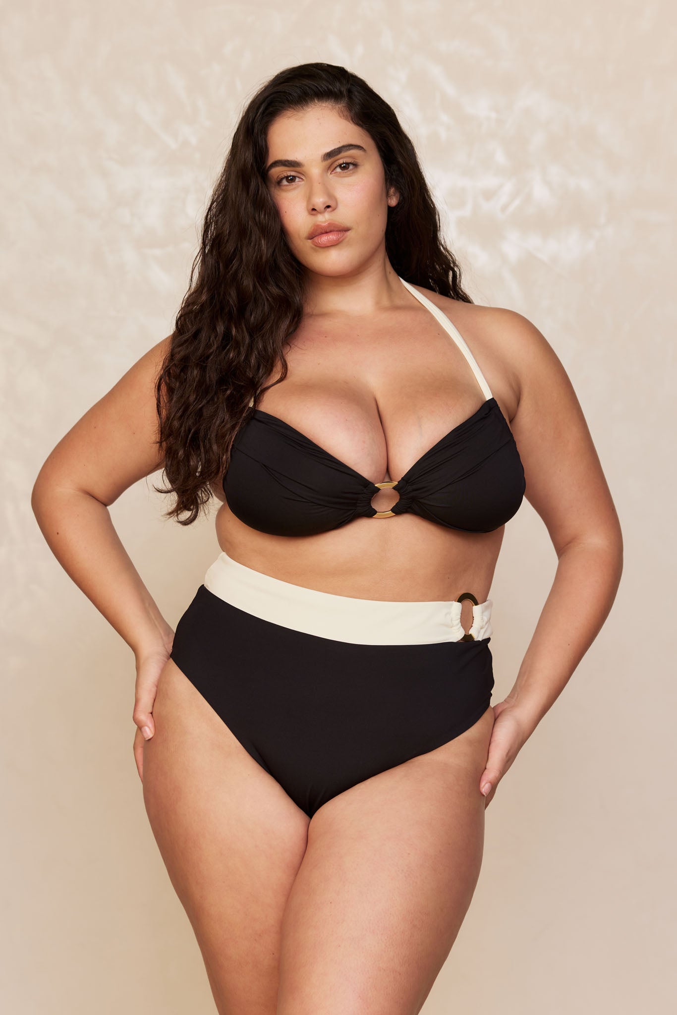 Plus-size model wearing a black high‑waist bikini with cream waistband and a black halter top with a front ring, hands on hips.