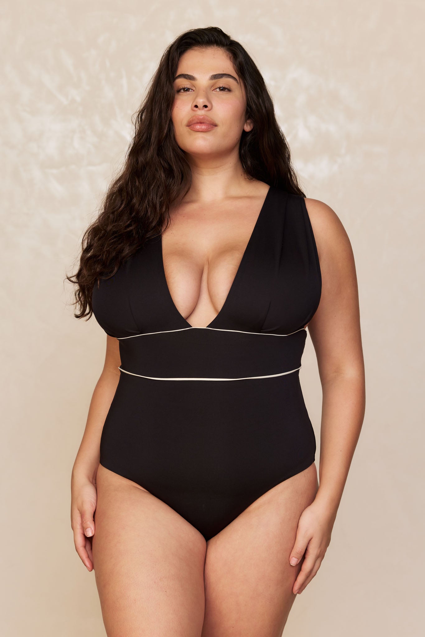Plus-size model in black one-piece with deep V neckline and thin white piping at underbust and waist, neutral background