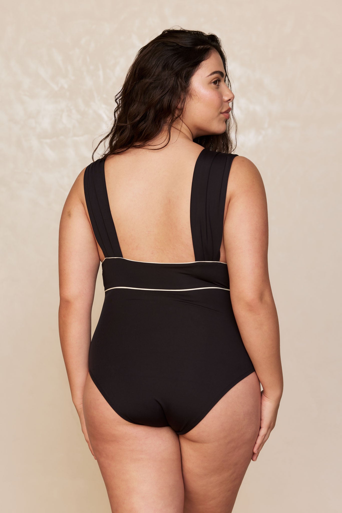 Plus-size woman seen from back wearing black one-piece swimsuit with wide shoulder straps and thin white waist piping