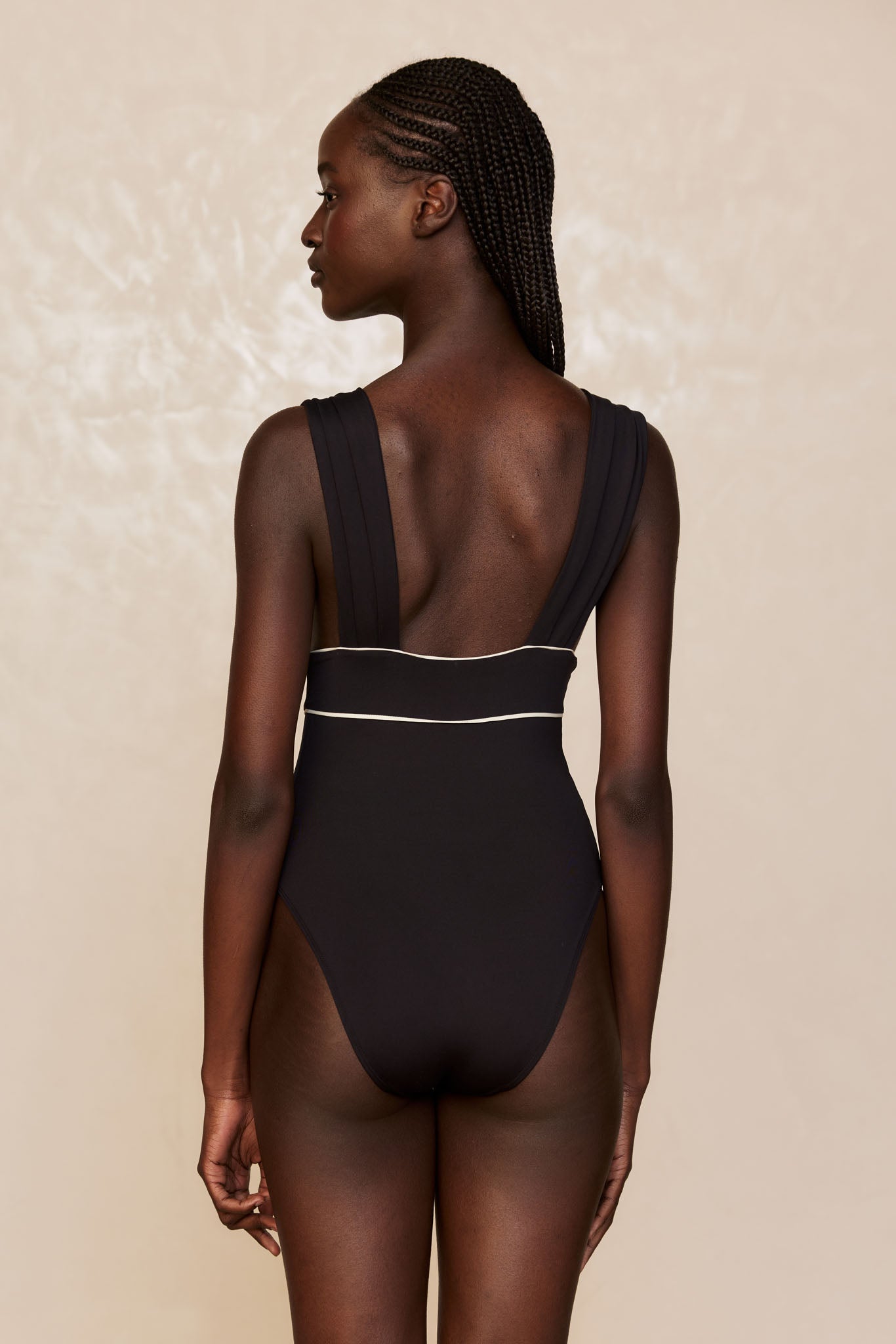 Back view of a woman with braided hair wearing a black one-piece swimsuit with wide straps and white piping at the waist
