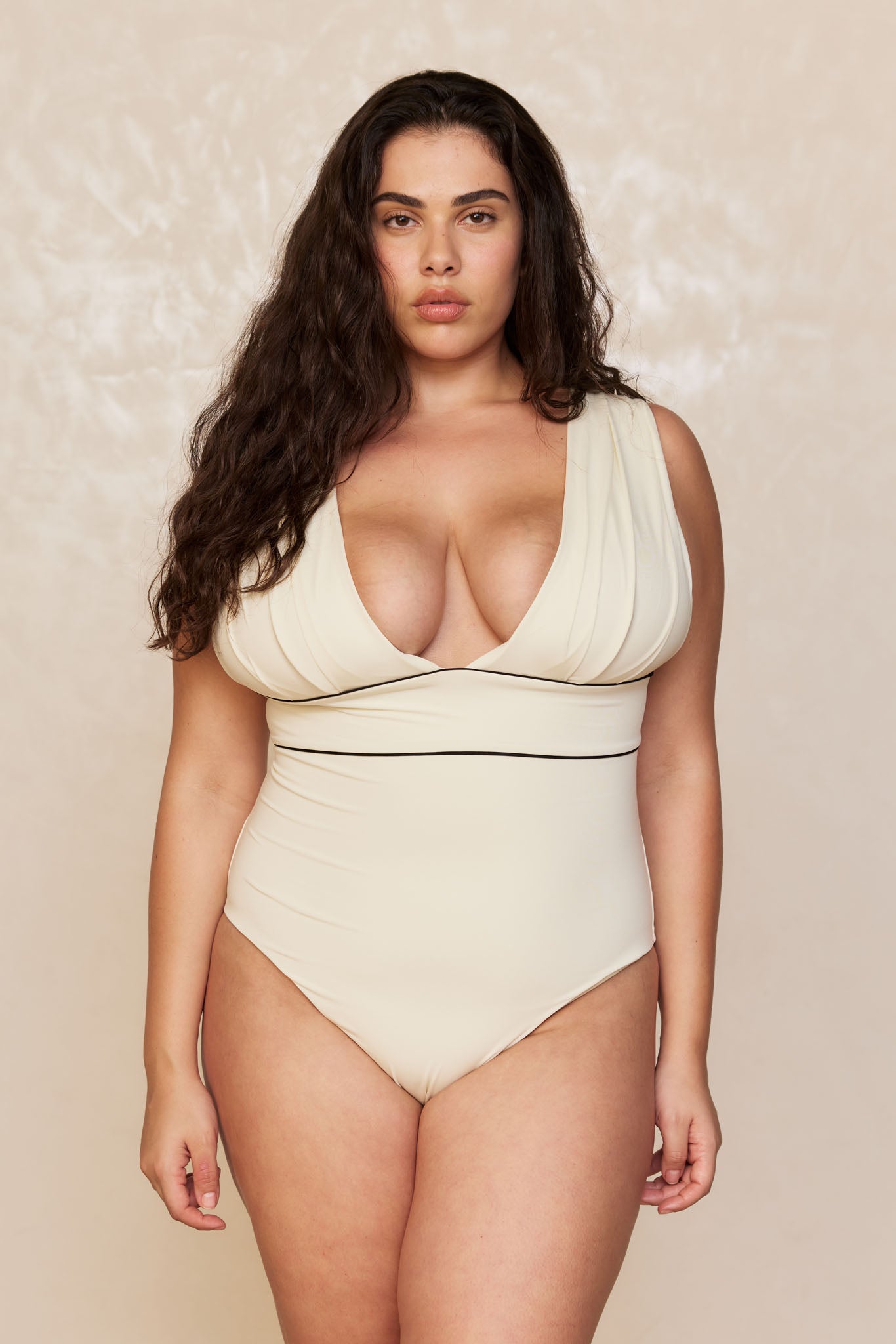 Plus-size woman with long dark wavy hair wearing a cream deep-V one-piece swimsuit, facing forward against neutral backdrop.