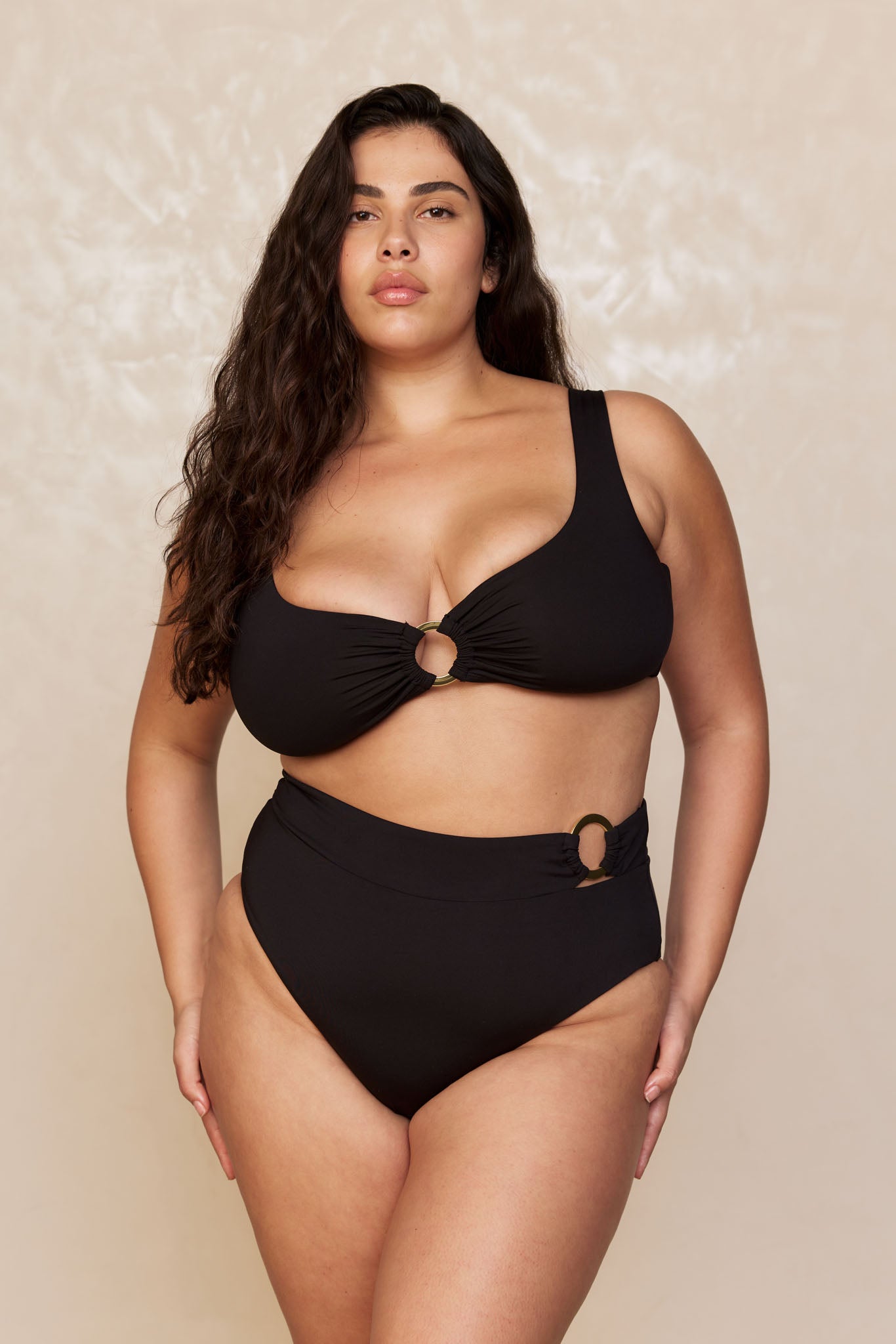 Plus-size woman wearing a black high-waist bikini with two ring accents, standing against a neutral textured backdrop.