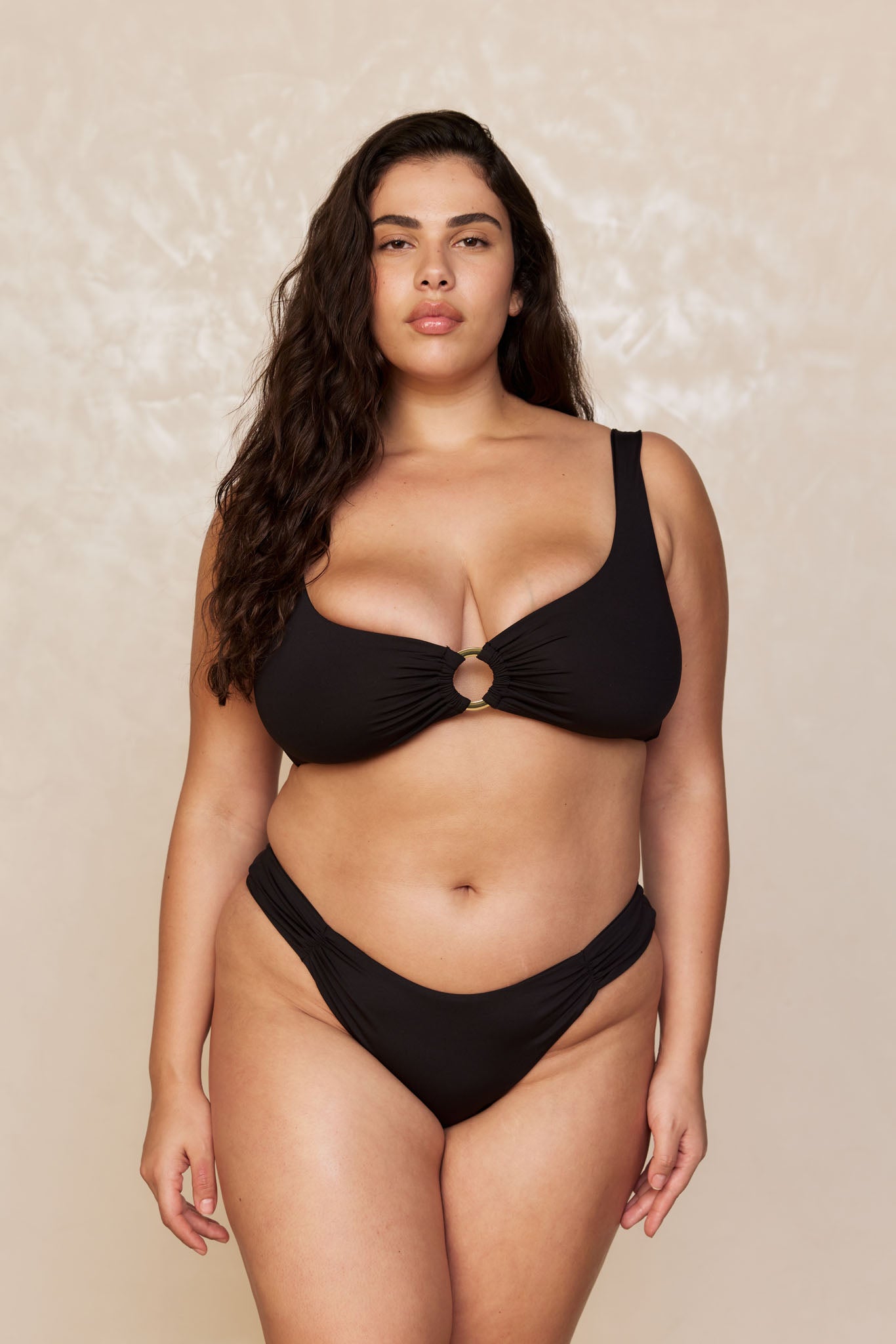Plus-size woman wearing black two-piece bikini with center ring detail, standing facing camera against beige backdrop.