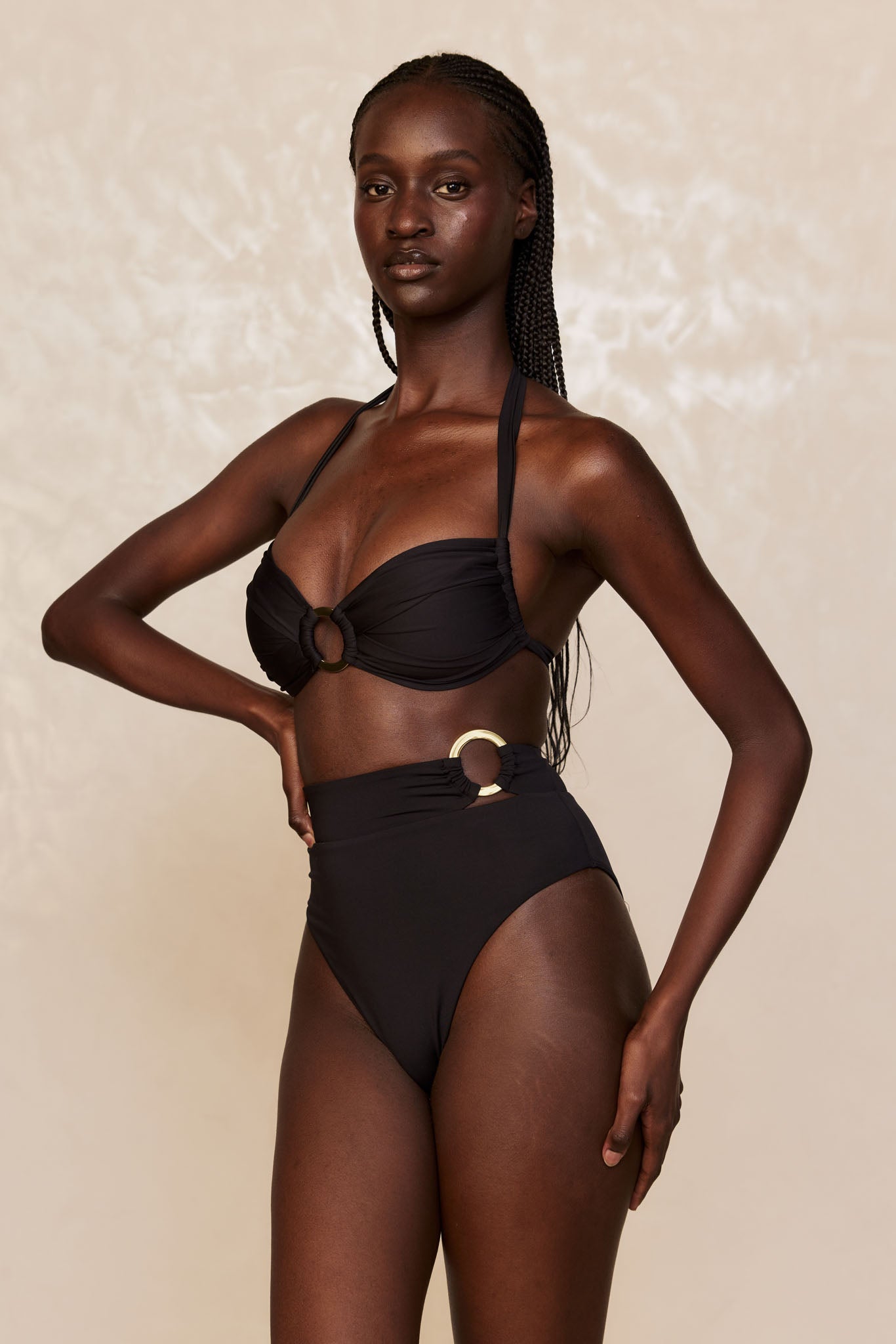 Female model wearing high-waist black bikini with gold rings, standing with one hand on hip against neutral backdrop.