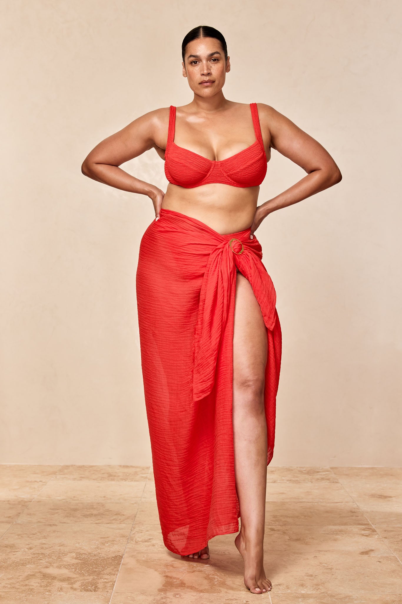 Plus-size woman wearing red bikini top and matching sarong with thigh-high slit, standing barefoot with hands on hips against neutral backdrop.