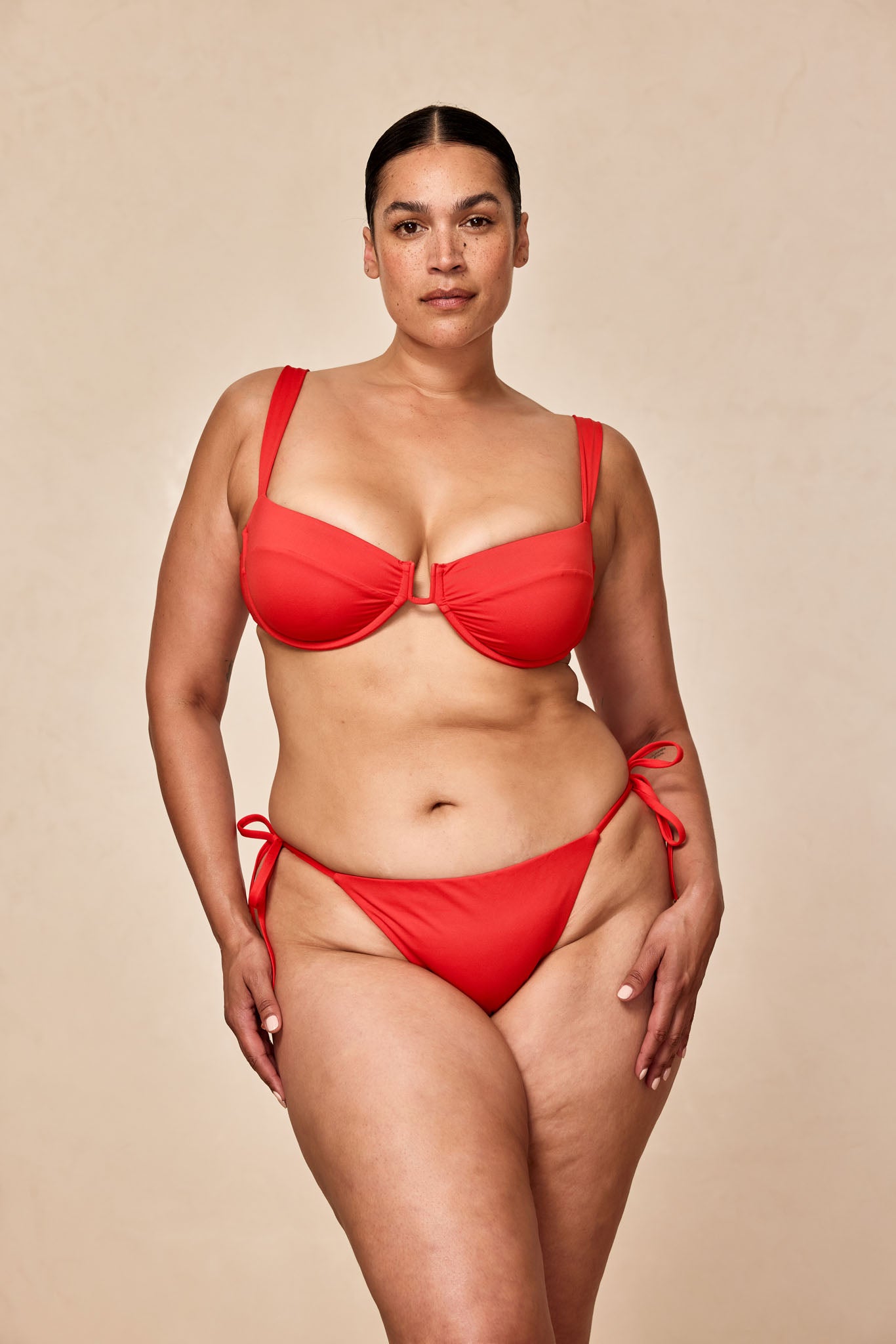 Plus-size woman standing in a red two-piece bikini against a neutral background, facing camera with hands at sides.