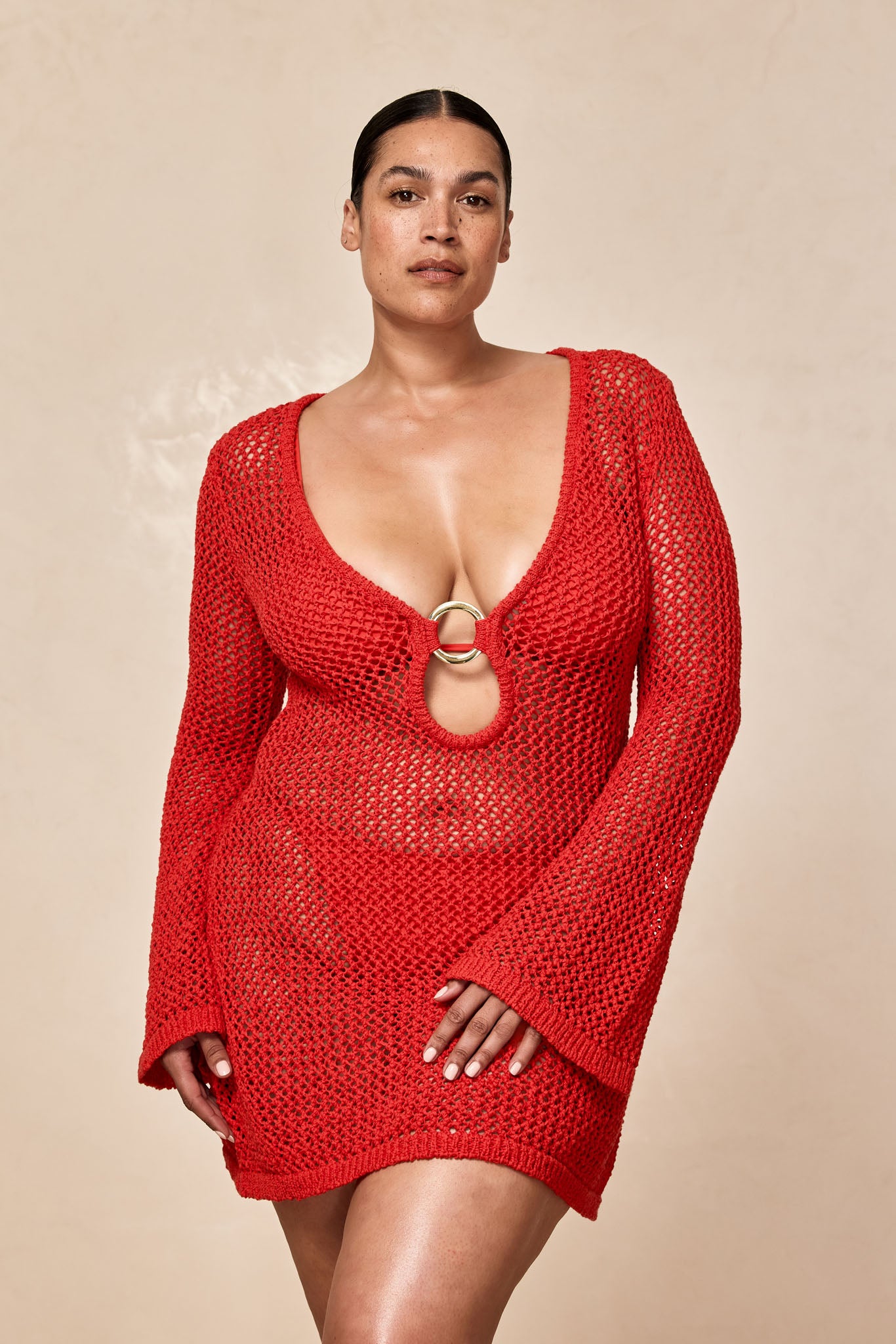 Model wearing a red open-knit, long-sleeve mini dress with deep scoop neckline and central gold ring at chest.