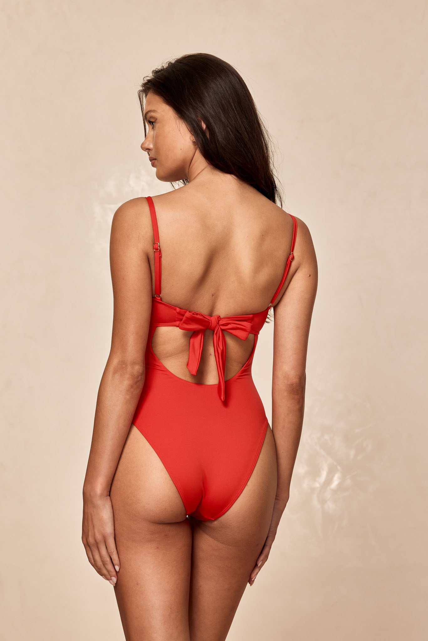 Back view of model wearing a red one-piece swimsuit with tied bow at back and adjustable shoulder straps