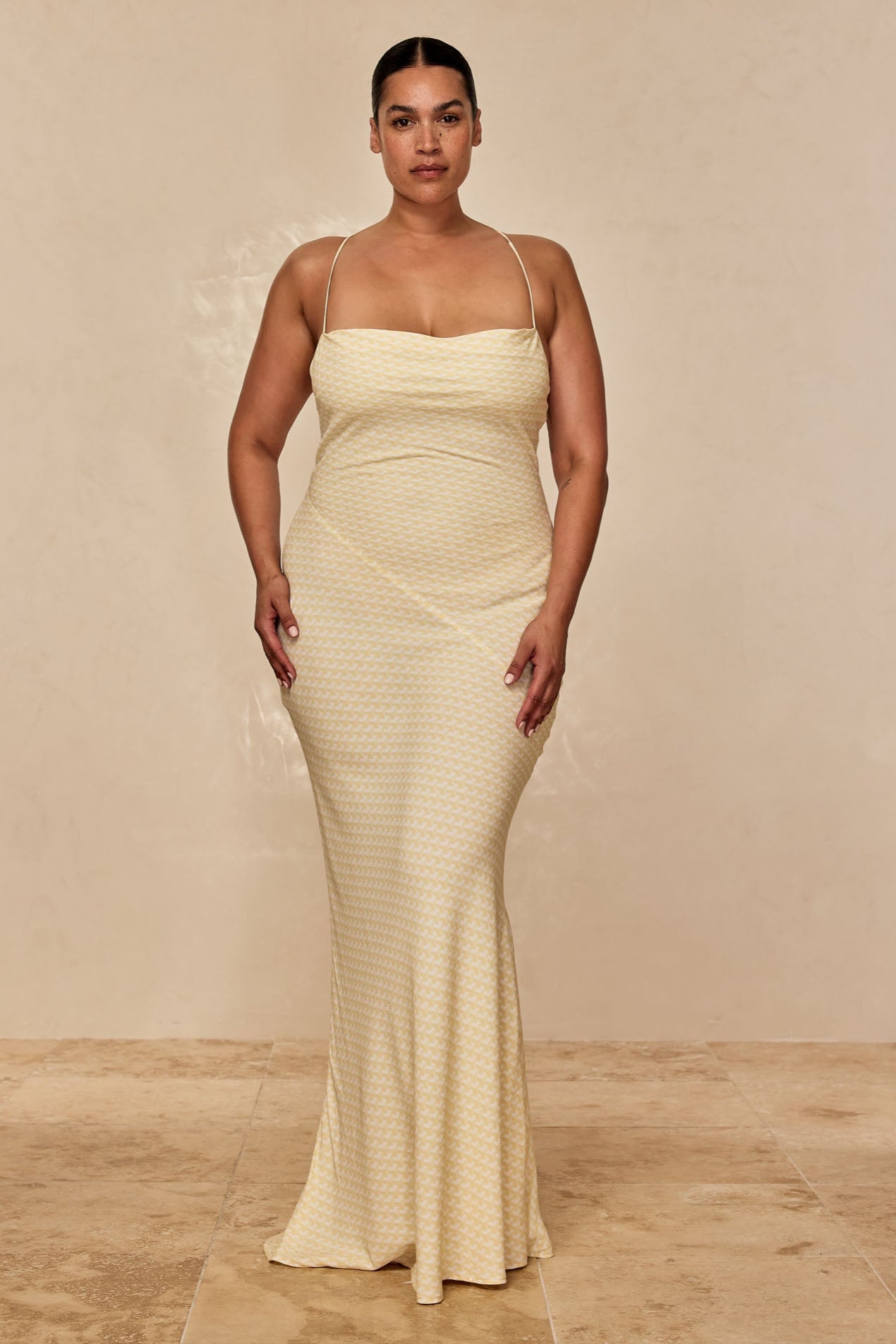 Model wearing a floor-length pale-yellow patterned slip dress with thin straps, standing against a neutral beige backdrop.