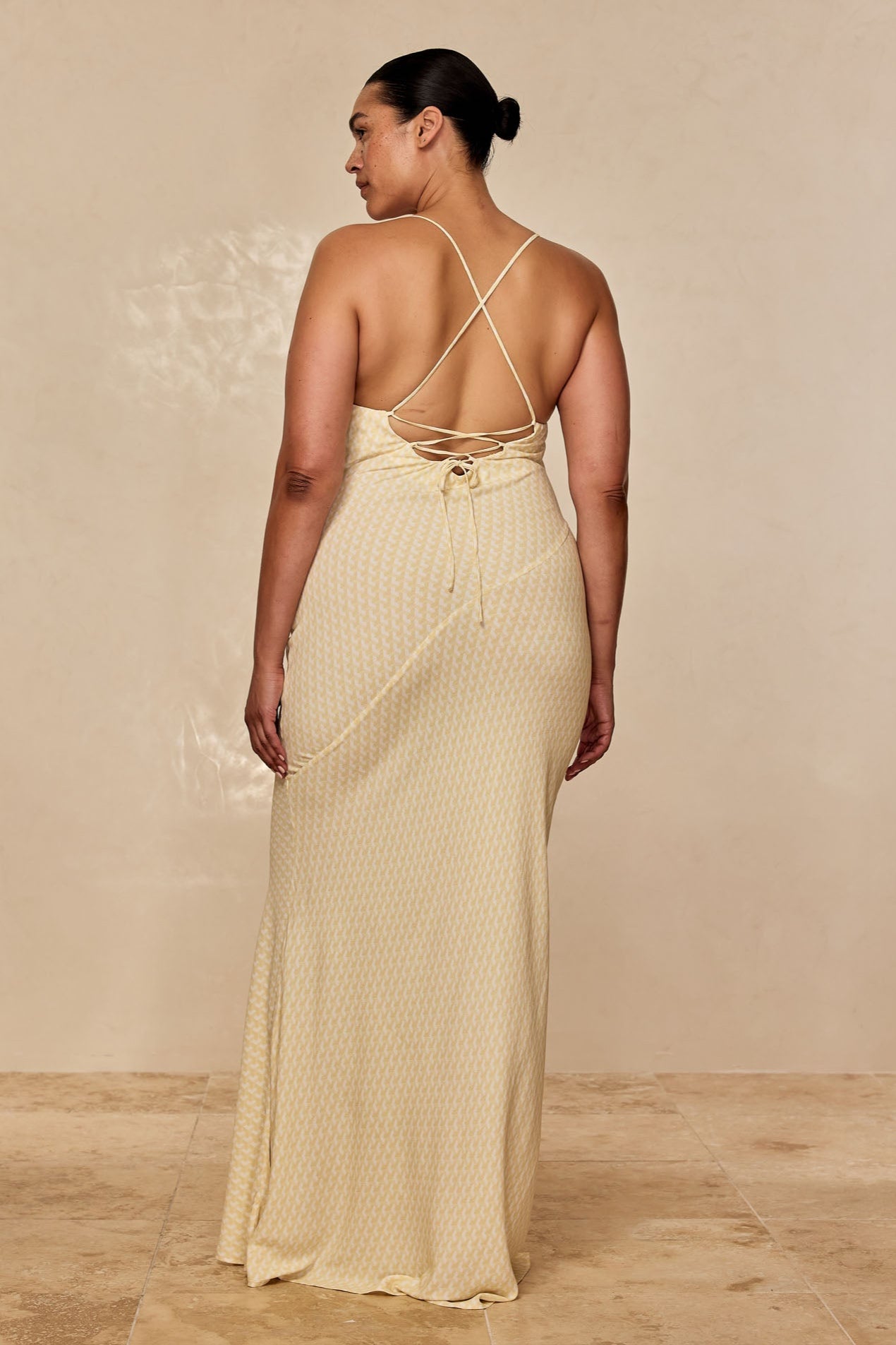 Woman seen from back wearing pale yellow patterned maxi dress with crisscross straps and tied back detail.