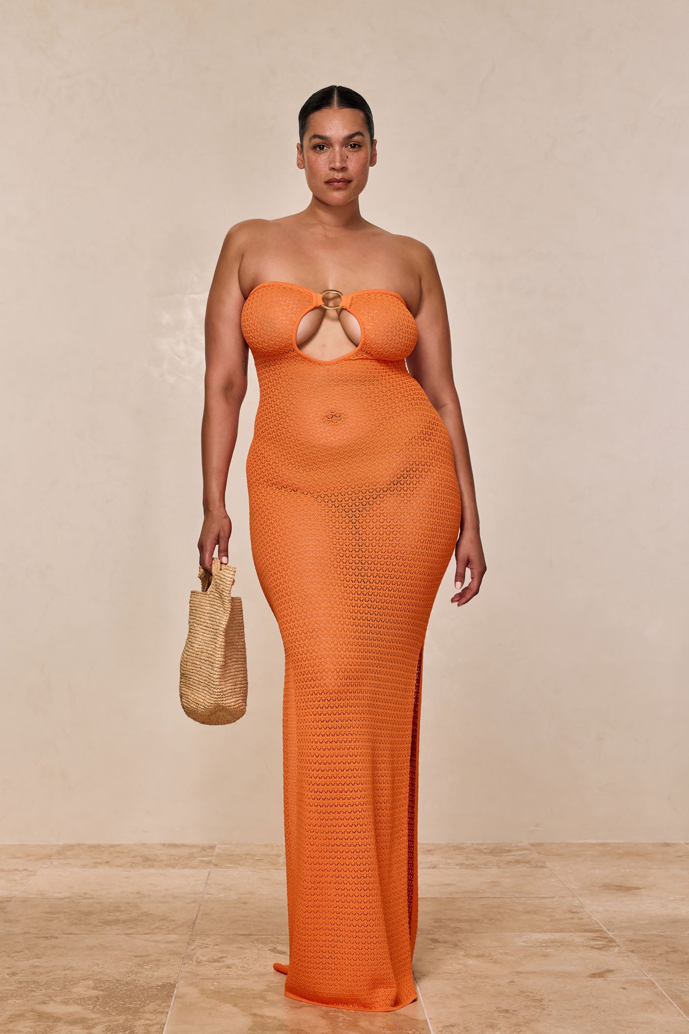 Plus-size woman wearing strapless orange crochet maxi dress with front ring cutout, holding small woven tote.