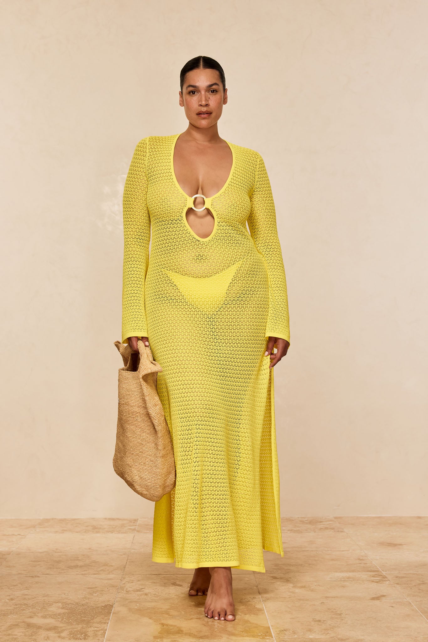 Plus-size model in yellow crocheted long-sleeve maxi dress with front ring cutout, carrying a straw tote.