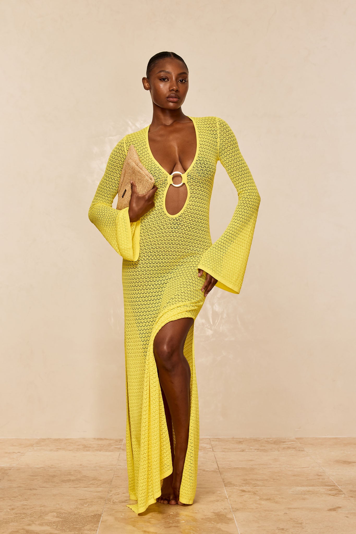 Model in yellow crochet maxi dress with front ring cutout and thigh-high slit, holding a straw clutch