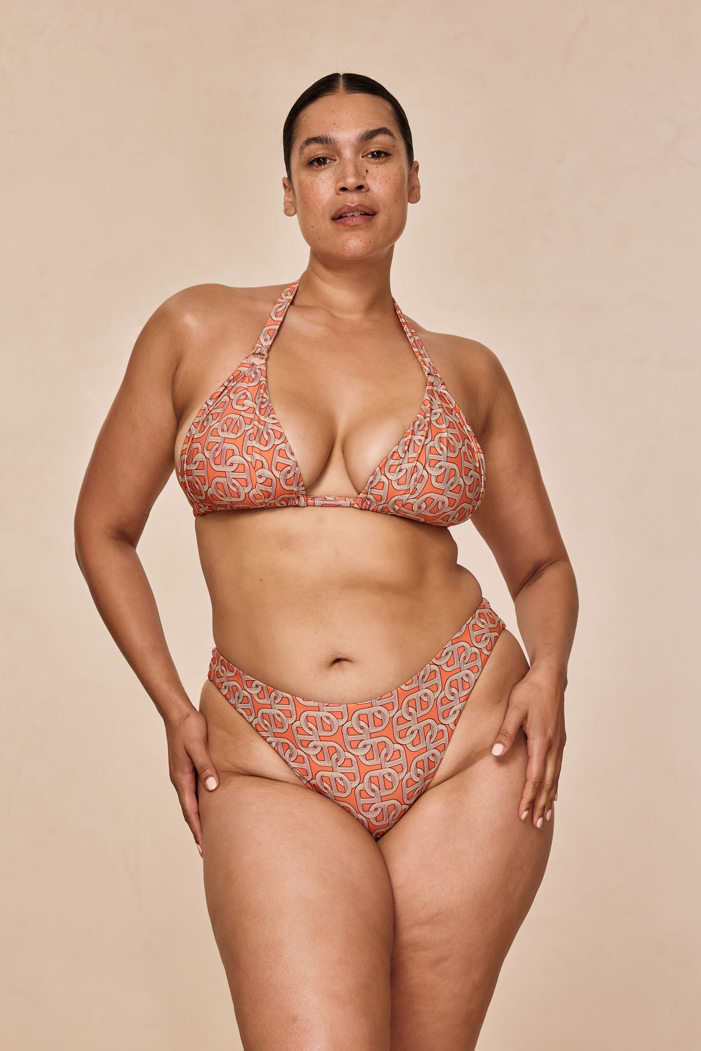 Model wearing orange patterned triangle bikini, standing with hands at hips against beige background.