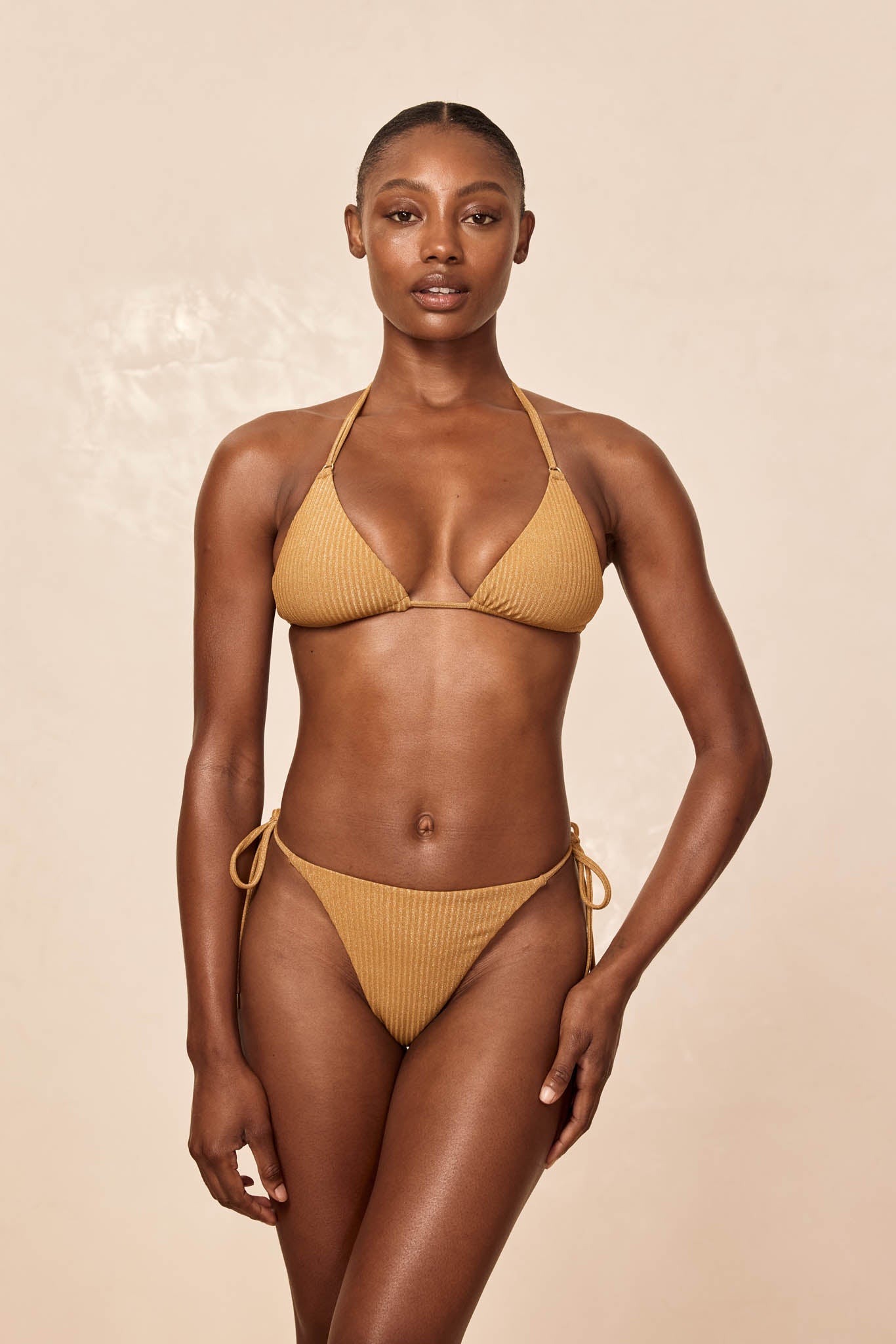 Front-facing female model wearing a mustard ribbed triangle bikini with tie-side bottoms on a beige background.