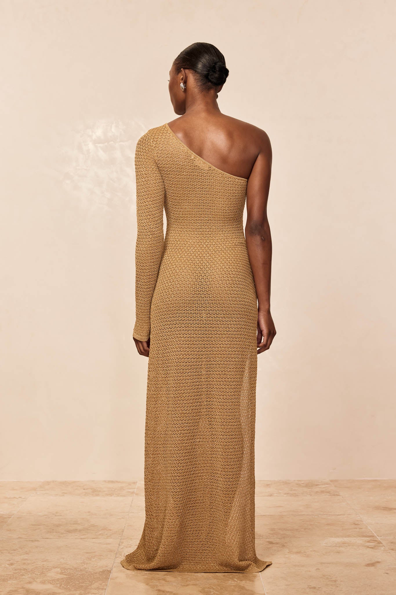 Woman viewed from back wearing a one-shoulder, long-sleeve knit gold gown with low bun hairstyle against a neutral background.
