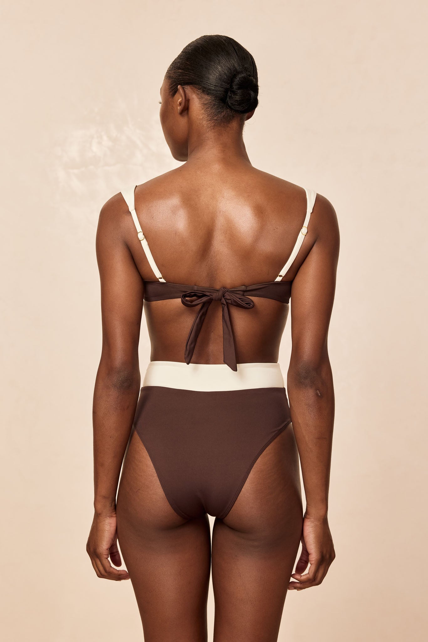 Female model shown from the back wearing a high-waisted brown and cream bikini with a tied back strap and low bun.